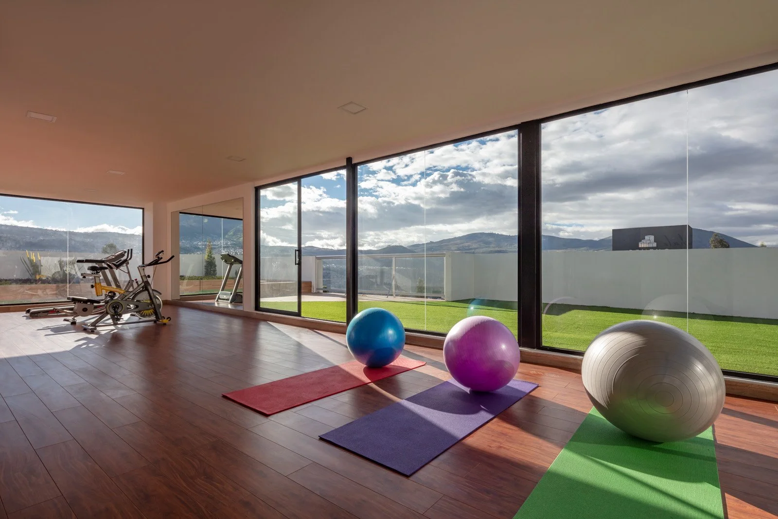 Sala de ejercicio con balones de yoga, tapetes y equipo de cardio, con vista a un jardín y montañas a través de ventanas grandes.