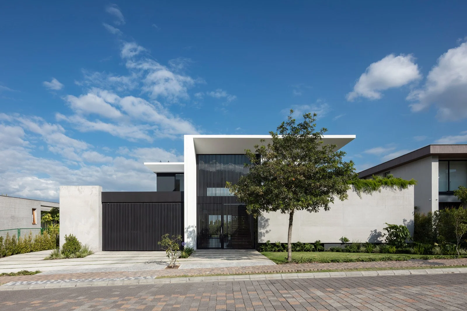 Casa moderna con paredes blancas y estructura de metal negro, rodeada de jardín y árboles, bajo un cielo azul con nubes.