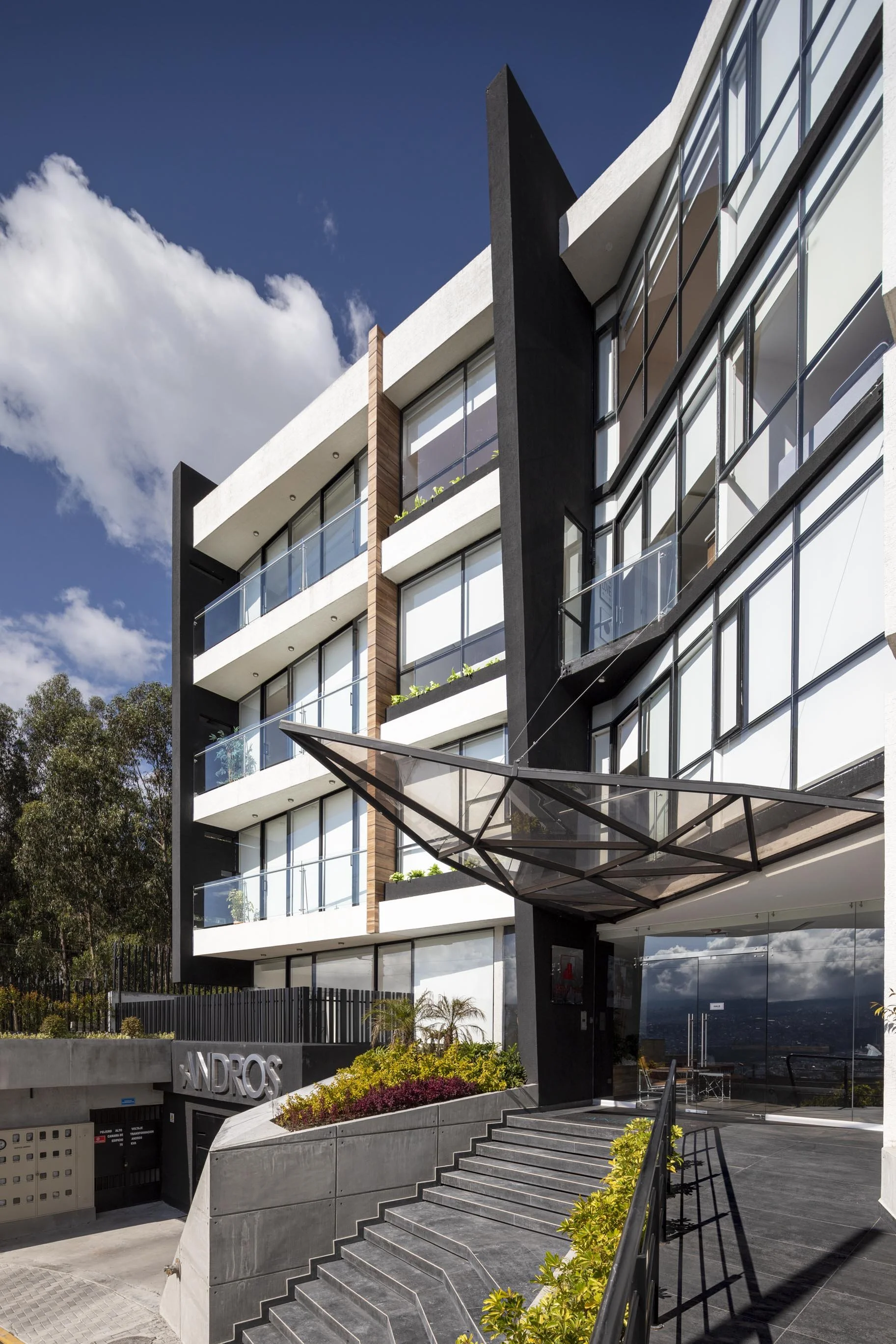 Edificio moderno con ventanas de cristales, balcones y una entrada con escaleras y pupitres de plantas y una superficie de vidrio en la parte superior, con el signo 'ANDROS' en la pared de la entrada.
