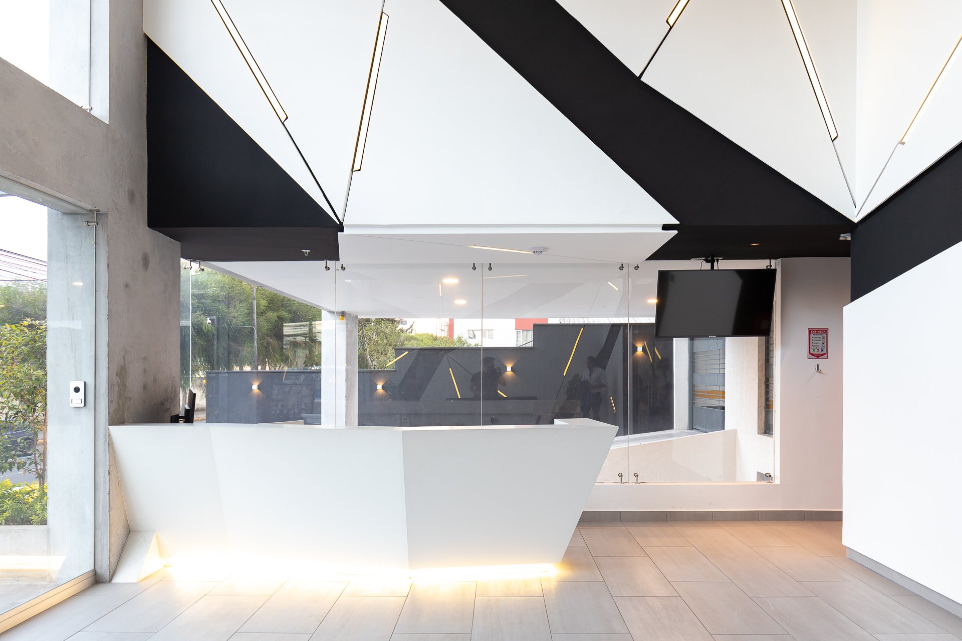 Recepción moderna con diseño minimalista en tonos blancos y negros, con un mostrador angular y paredes decoradas con líneas de luz LED, además de un televisor en la esquina y amplios ventanales que dejan entrar luz natural.