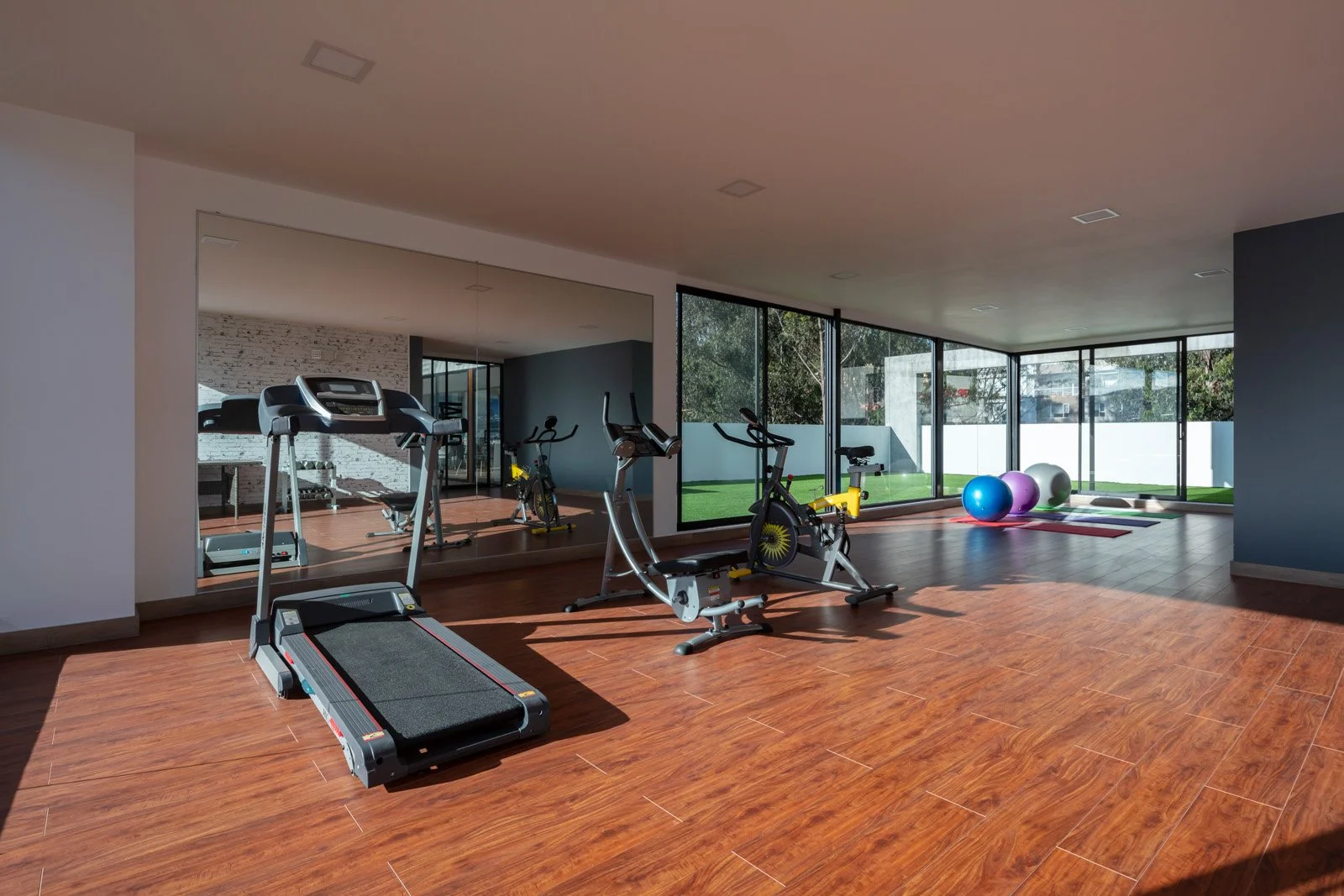 Sala de ejercicio con cinta de correr, bicicletas y peso libre, con ventanas grandes y piso de madera.