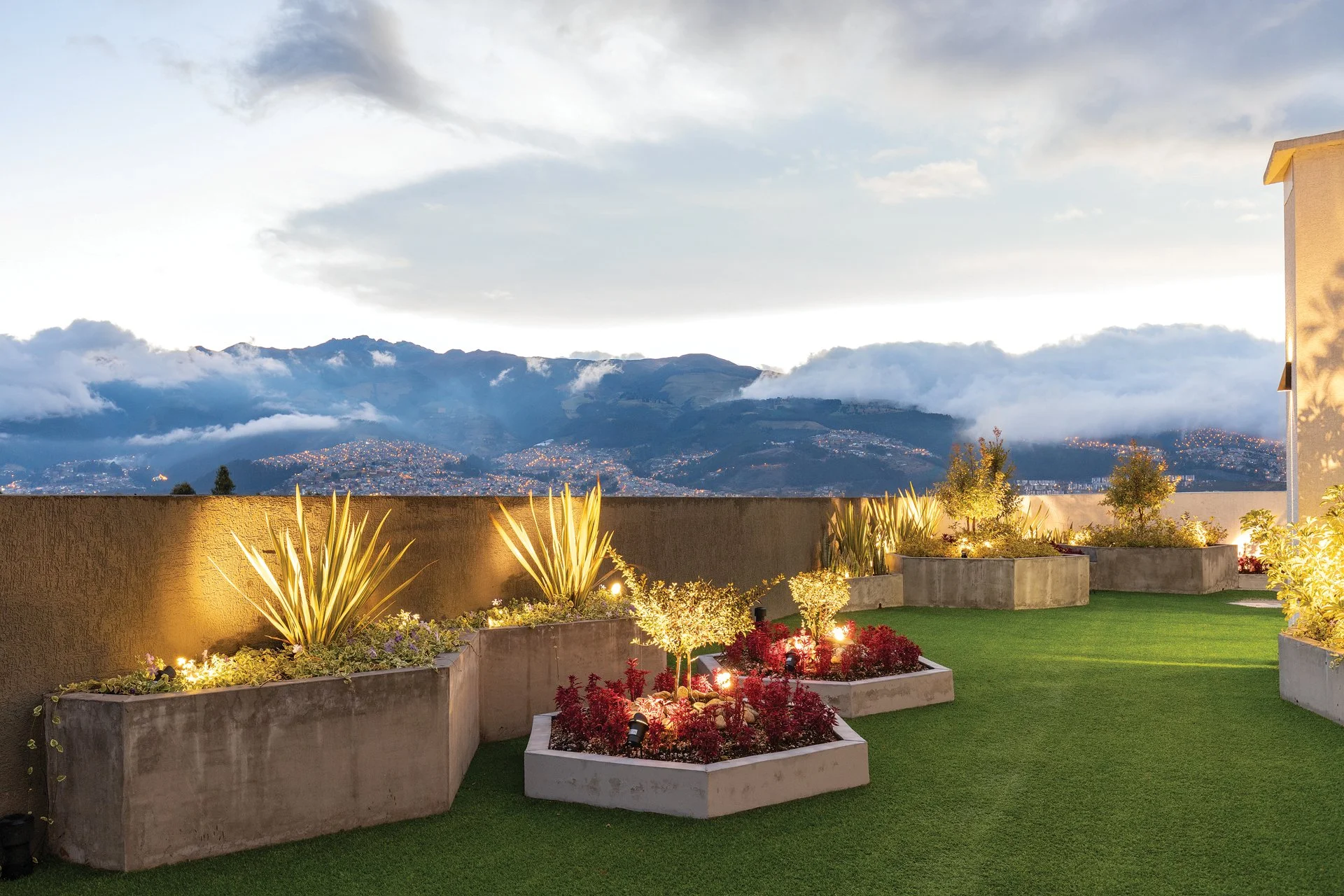 Terraza con jardineras de concreto llenas de plantas y árboles iluminados, con vista a una cadena montañosa con nubes y ciudad al fondo al atardecer.