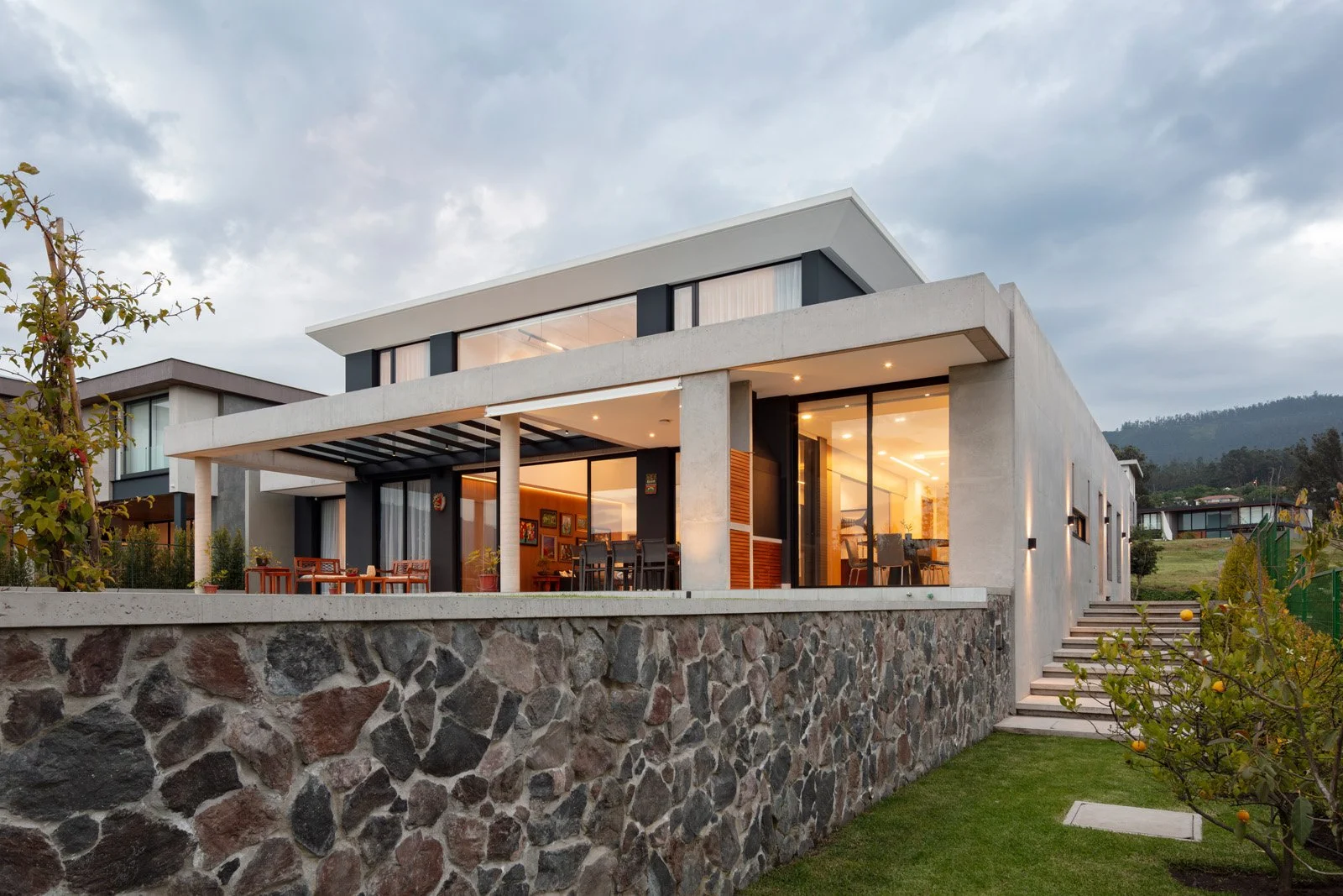 Casa moderna de dos pisos con fachada de concreto y piedra en un entorno rural, con jardines y montañas al fondo.