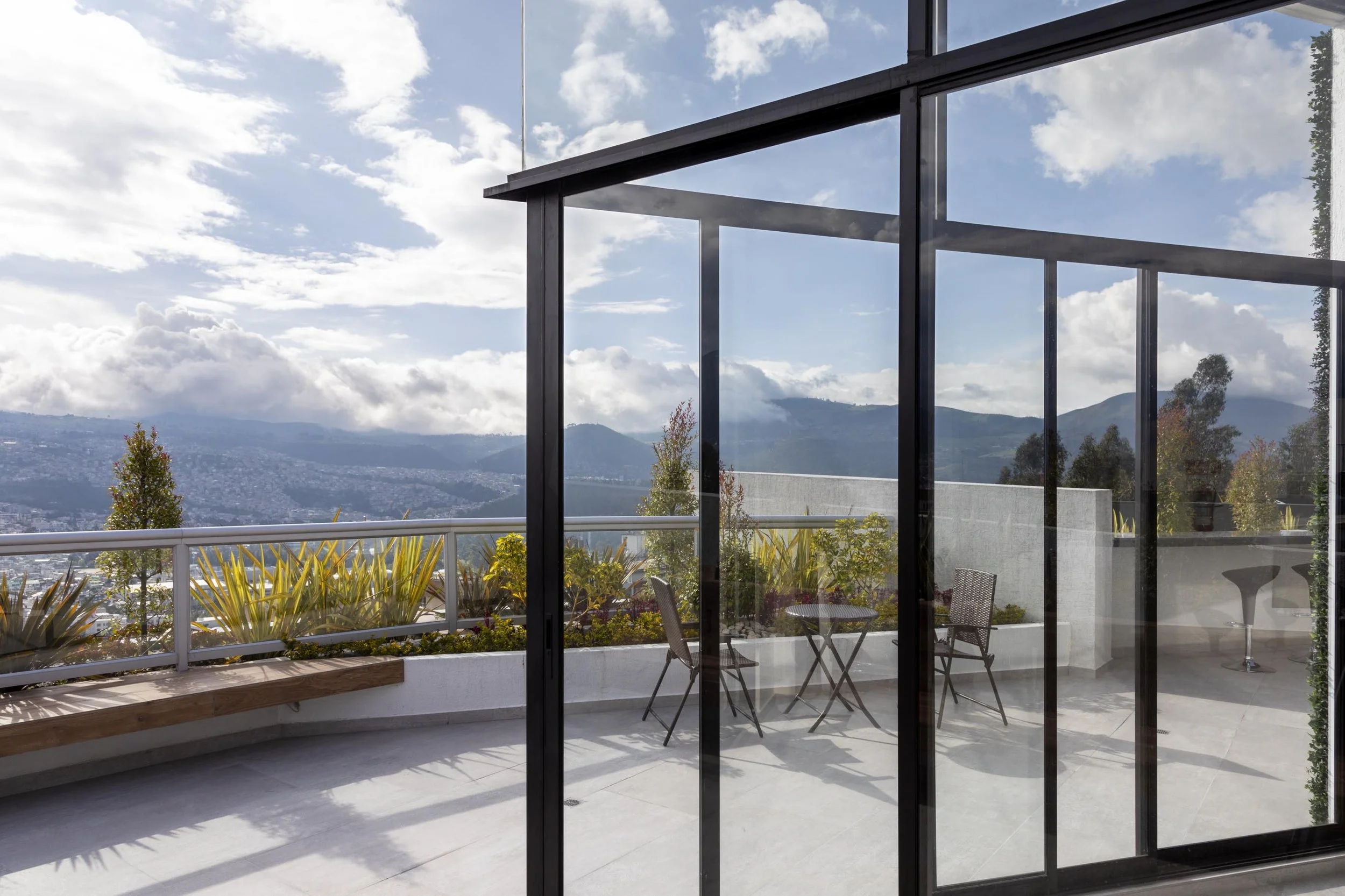Vista desde una terraza con muebles de jardín, plantas y montañas al fondo, vista a través de ventanas de cristal y una estructura de metal negro.