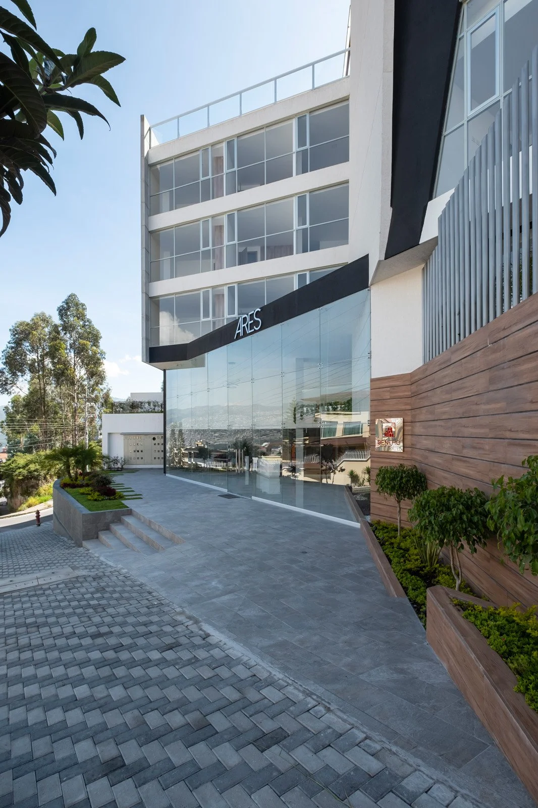 Edificio moderno con paredes de vidrio y señal que dice 'ARES' frente a la entrada, con plantas y escaleras en la acera.