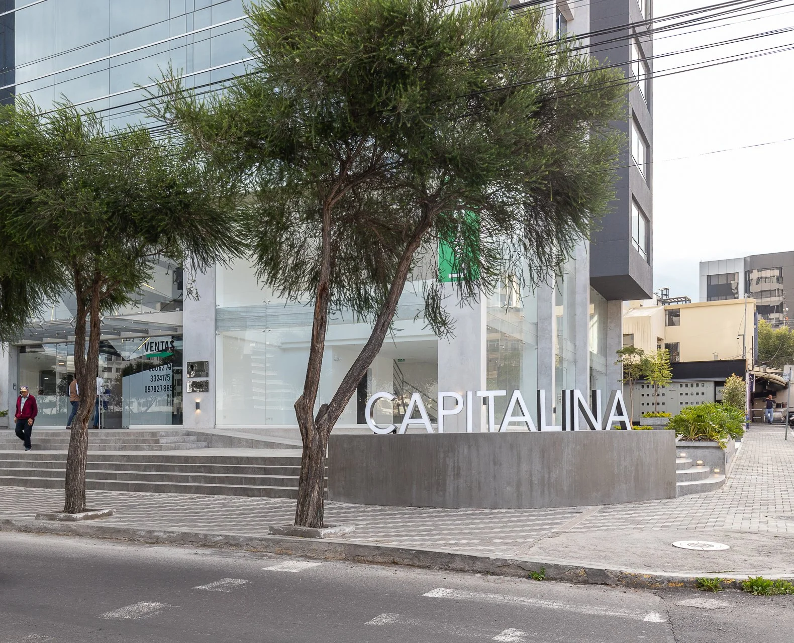 Fachada de un edificio moderno con la palabra 'CAPITALINA' en letras grandes, en la acera hay dos árboles y unas escaleras. En el fondo hay otras construcciones y un cielo nublado.