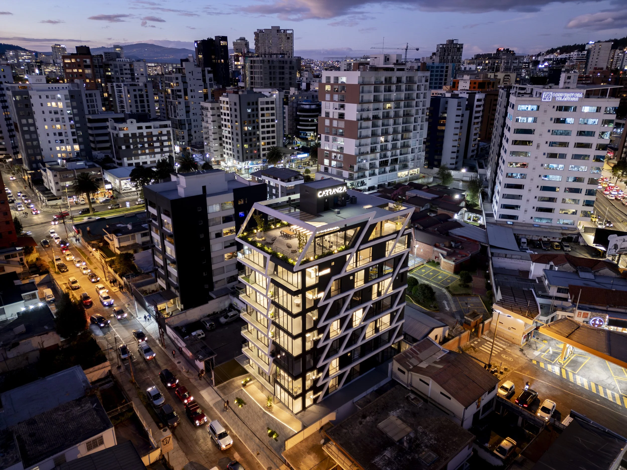 Vista aérea de una ciudad moderna con edificios altos y calles iluminadas al atardecer, resaltando un edificio con un diseño arquitectónico innovador y terraza en la azotea.