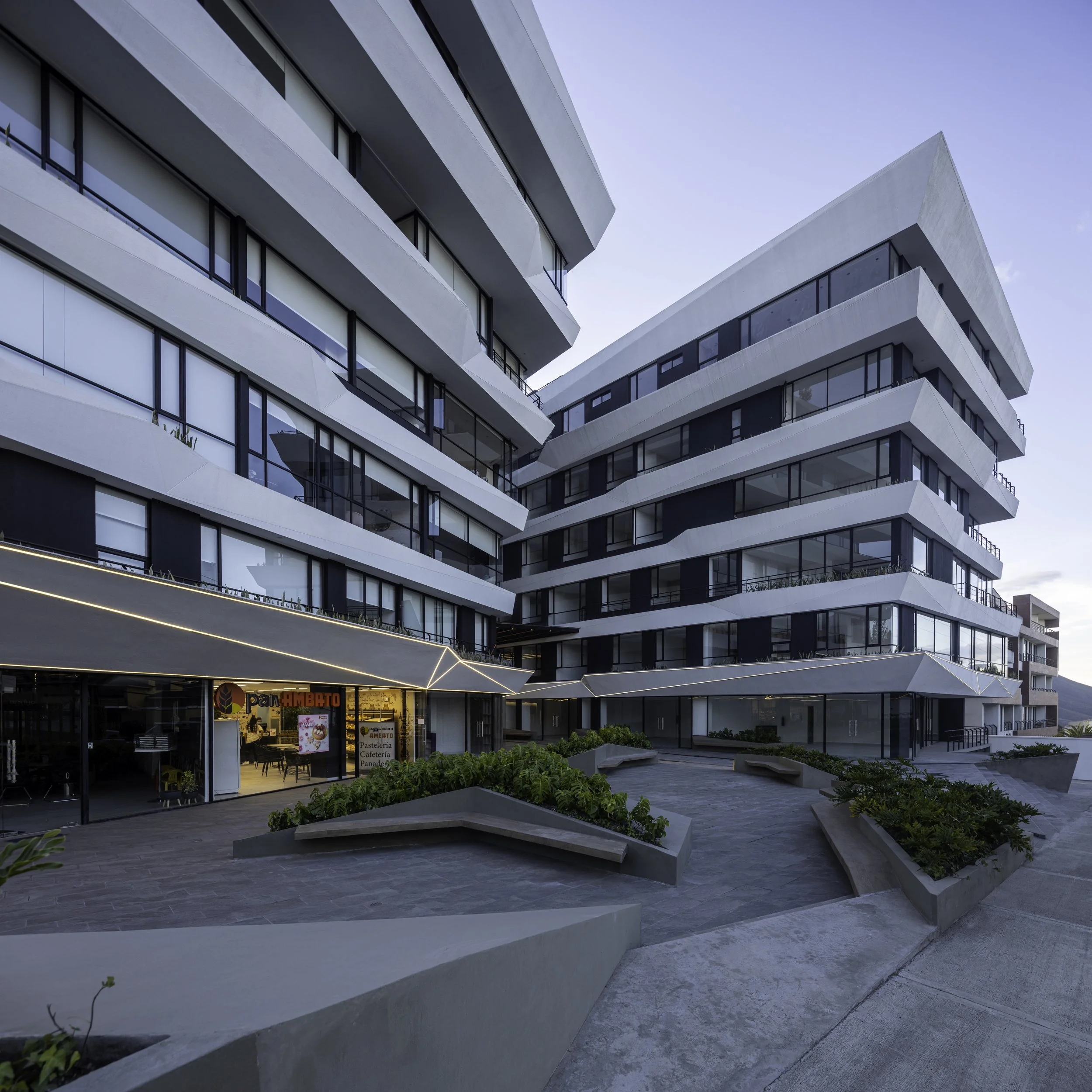 Edificio moderno de varias plantas con balcones y grandes ventanas, con un local de comida en la planta baja y áreas de jardinería y caminos en el área exterior.
