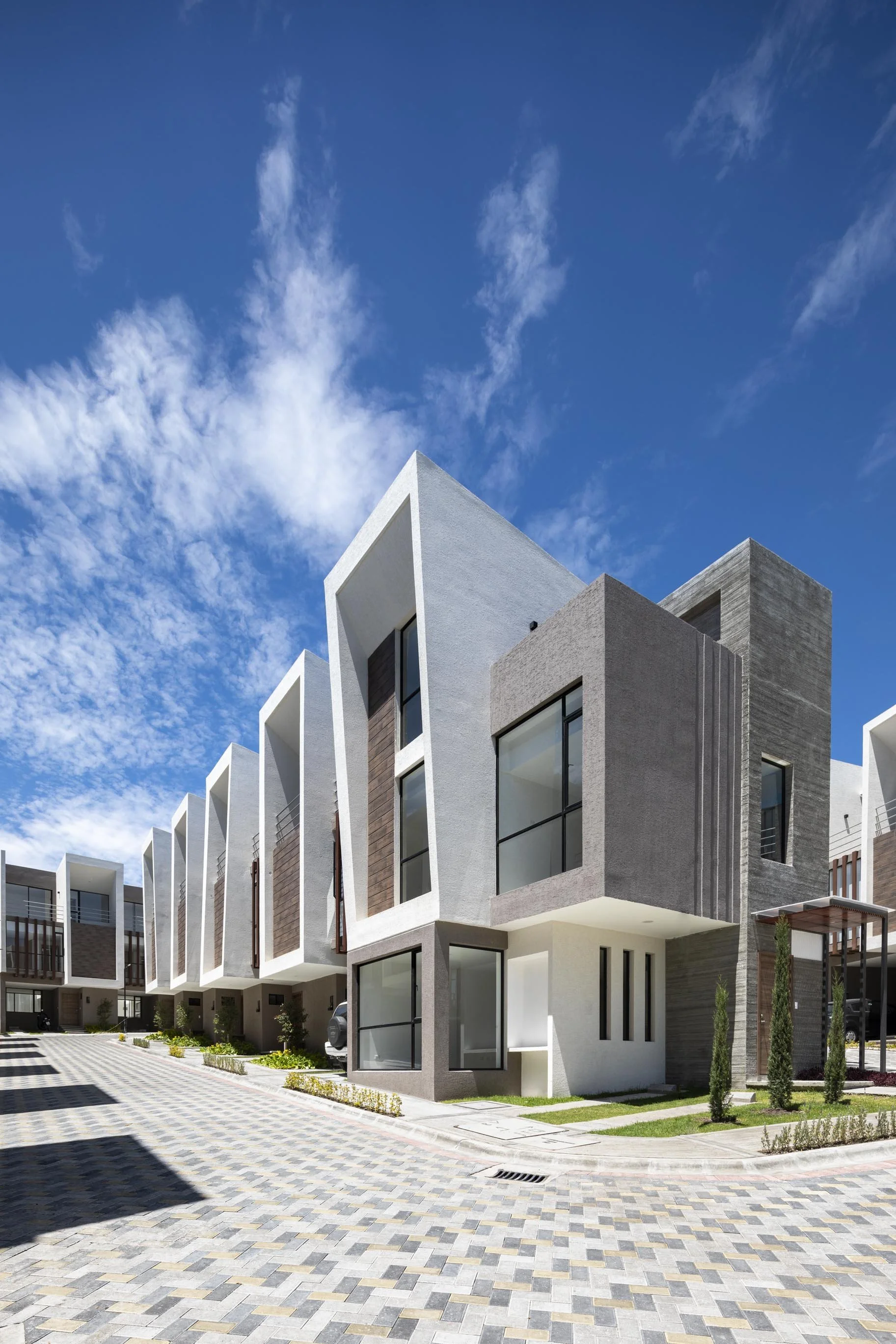 Conjunto de casas modernas con fachadas blancas, grises y de madera, con ventanas grandes en un día soleado y cielo con nubes