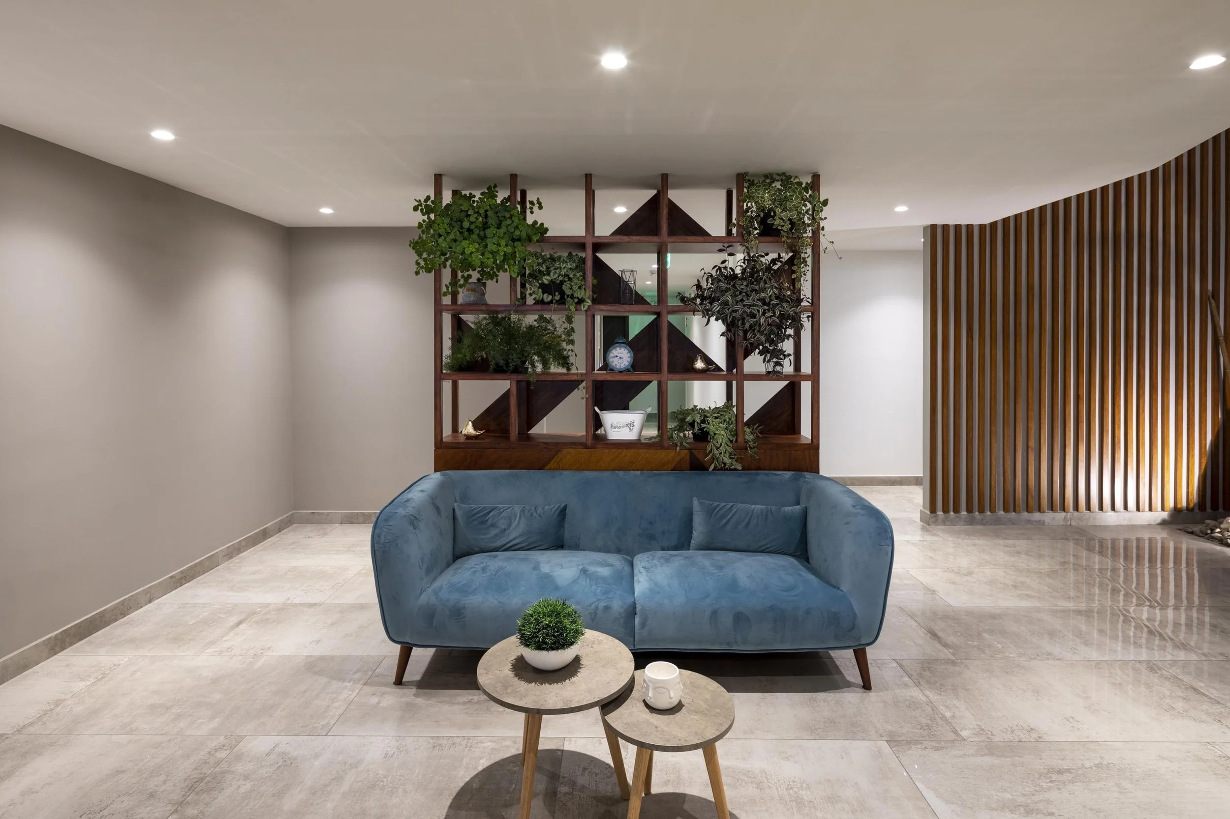 Sala moderna con sofá azul, mesas pequeñas, estantería de madera con plantas y objetos decorativos, paredes blancas y piso de baldosas beige.