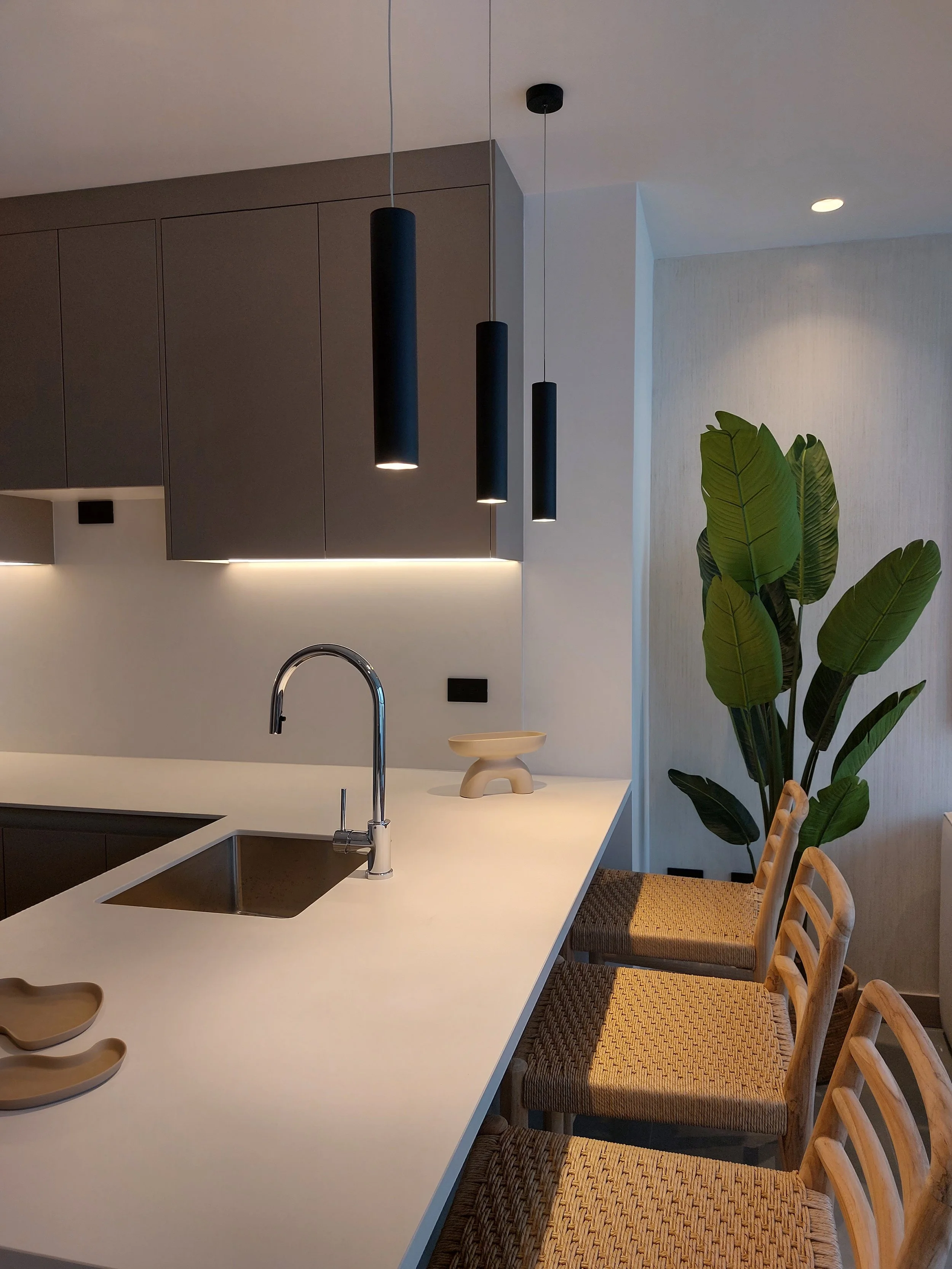 Cocina moderna con isla blanca, cuatro sillas de madera con asientos de fibra en la barra, lavaplatos con grifo de acero, plantas grandes en la esquina y lámparas negras colgantes.