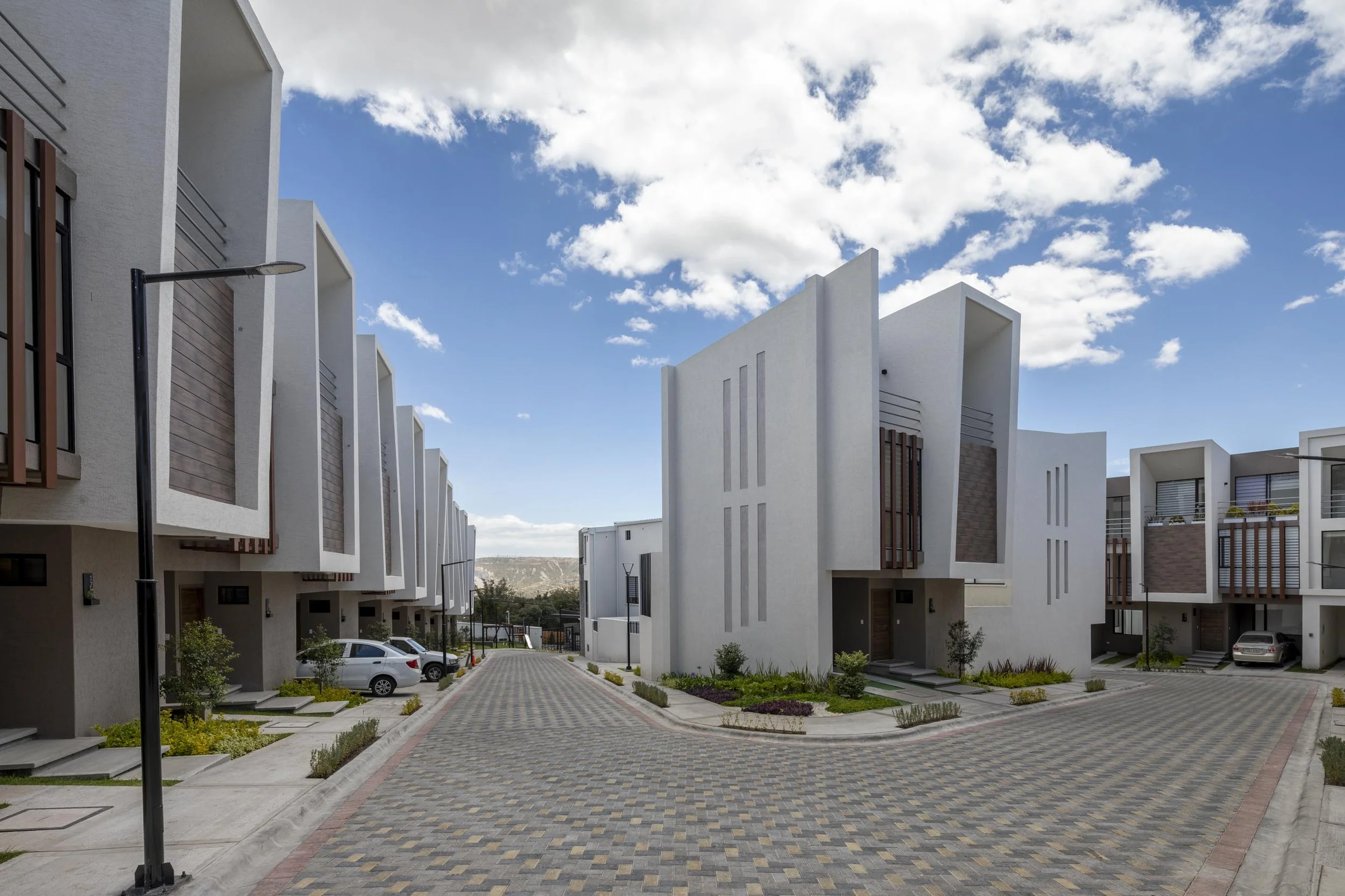 Conjunto habitacional de casas modernas blancas con estructuraciones de madera en las fachadas, áreas verdes, coches estacionados y calles de pavimento en un día soleado con cielo despejado y algunas nubes.