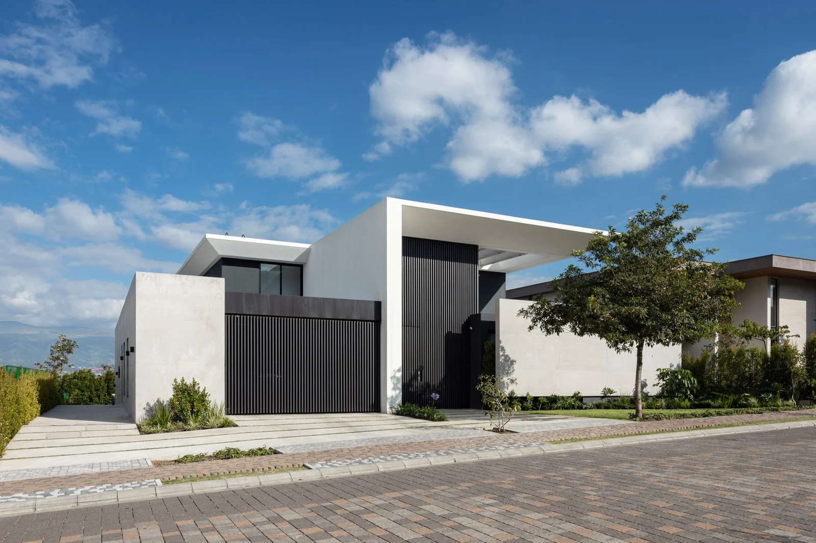 Casa moderna de diseño minimalista con muros blancos, puertas negras, y un jardín con árbol y plantas en un vecindario con un cielo azul y nubes.
