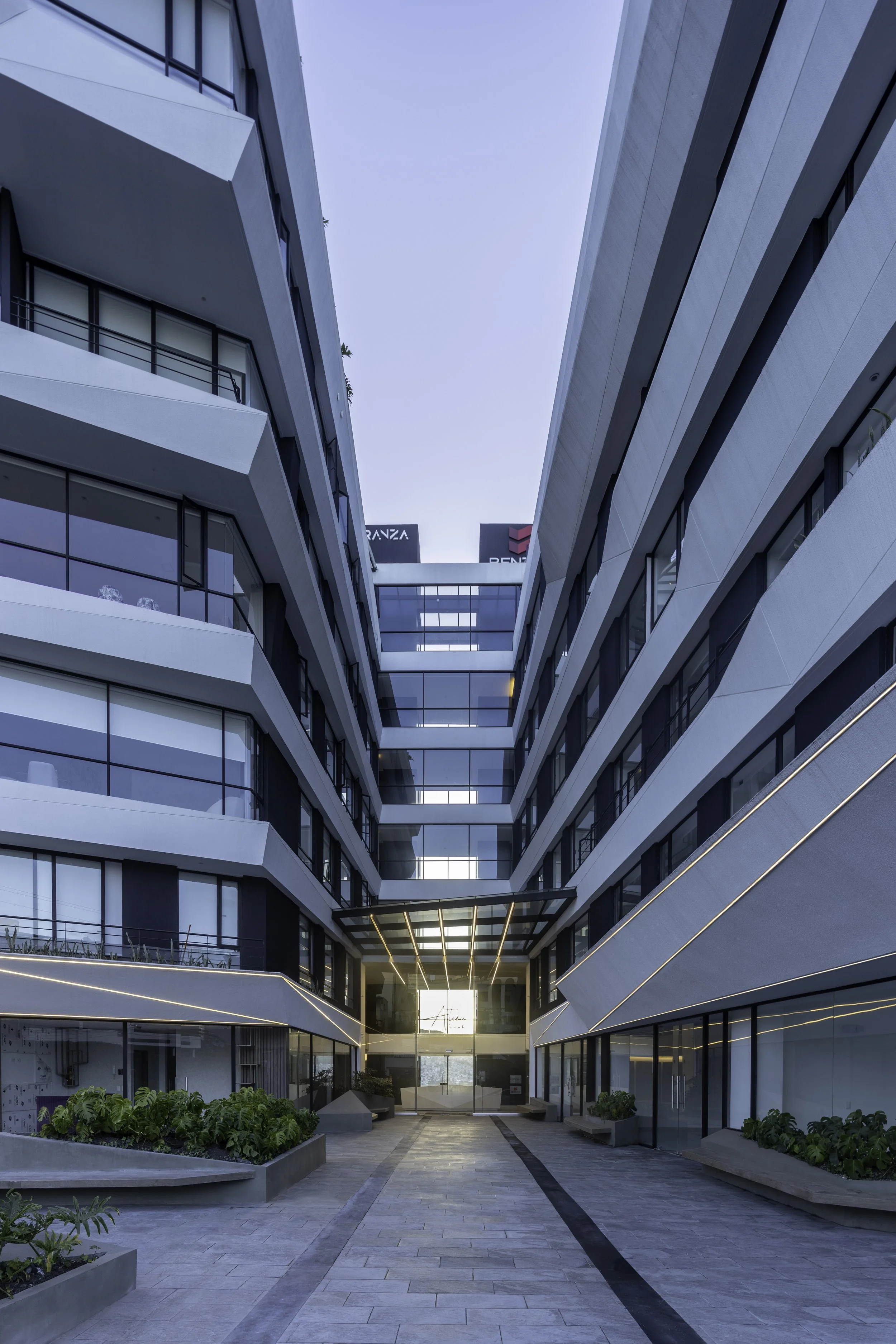 Vista de un edificio moderno y elegante con pasillo central al aire libre, plantas en jardineras y ventanas de vidrio que reflejan el cielo