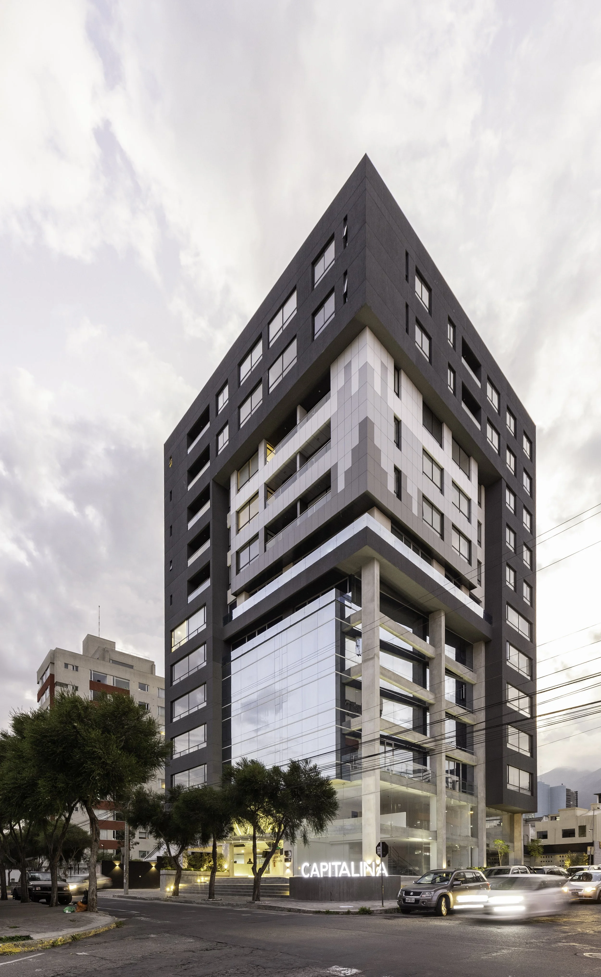 Edificio moderno de varios pisos en una ciudad, con fachada de vidrio y estructura de concreto, cerca de árboles y autos en la calle, con el letrero 'CAPITALINA' en la entrada.