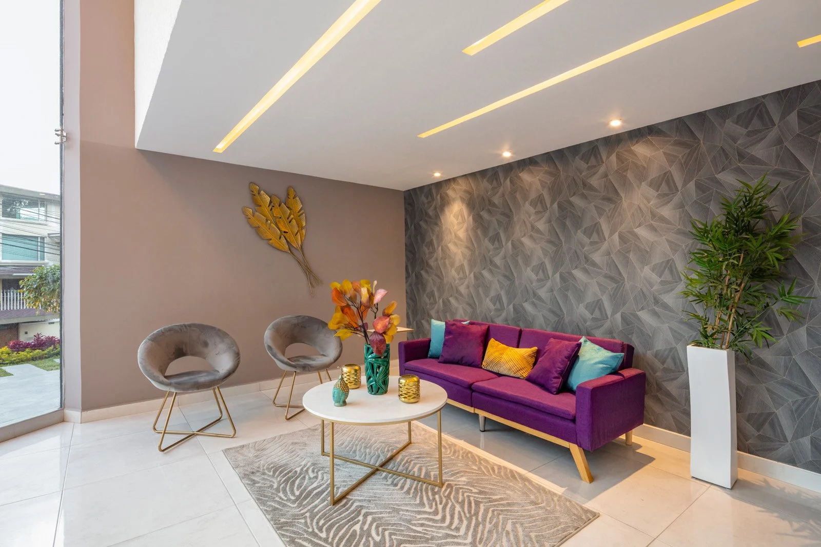 Sala de estar moderna con sofá violeta, dos sillas de terciopelo gris, mesa blanca con decoración floral y objetos decorativos dorados, pared gris con textura geométrica, una planta grande en una maceta blanca y detalles de iluminación en el techo.