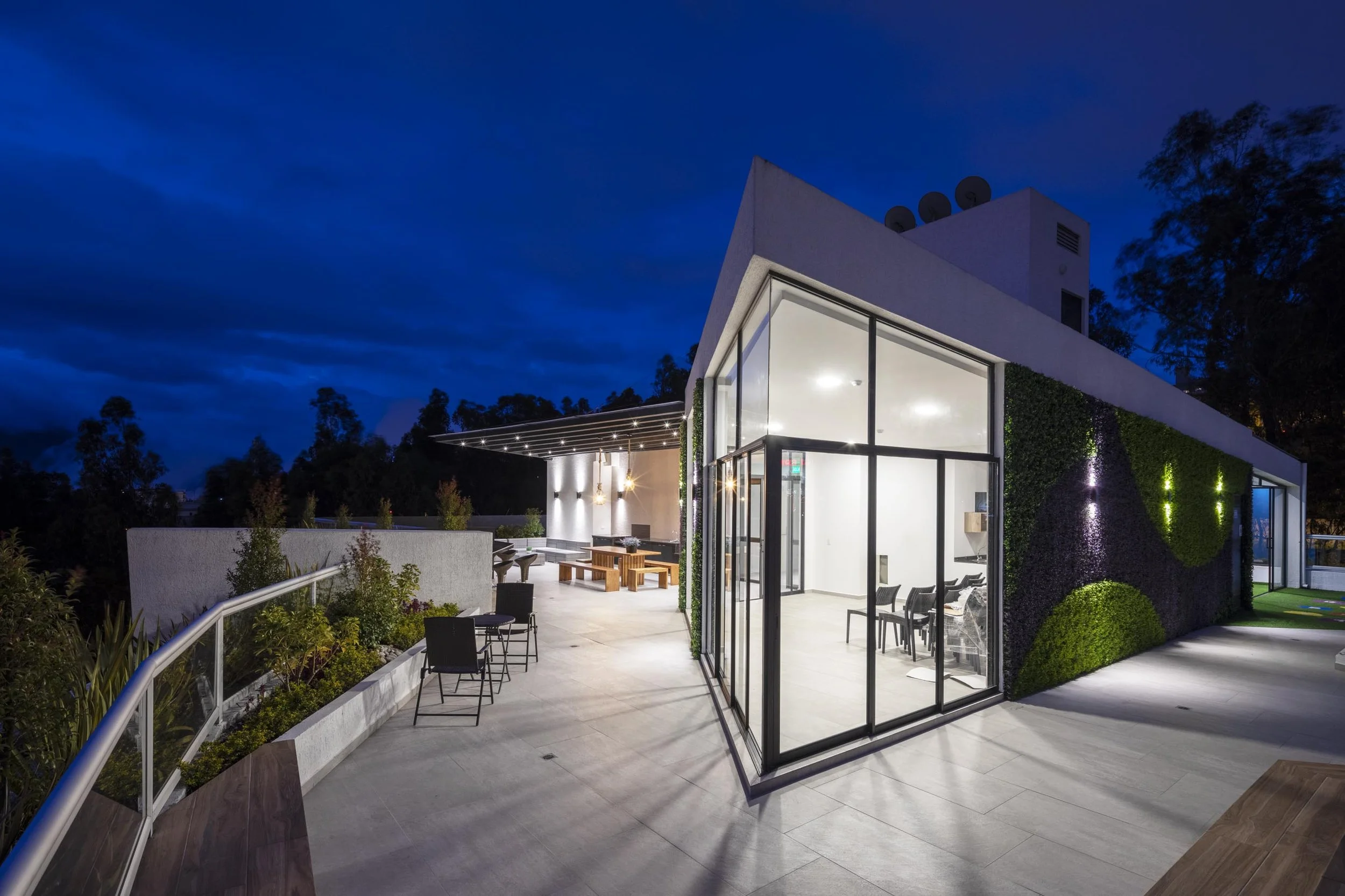 Terraza moderna en un edificio con paredes de cristal y vegetación decorativa, iluminada por luces de noche, en un entorno nocturno con cielo oscuro y árboles en el fondo.
