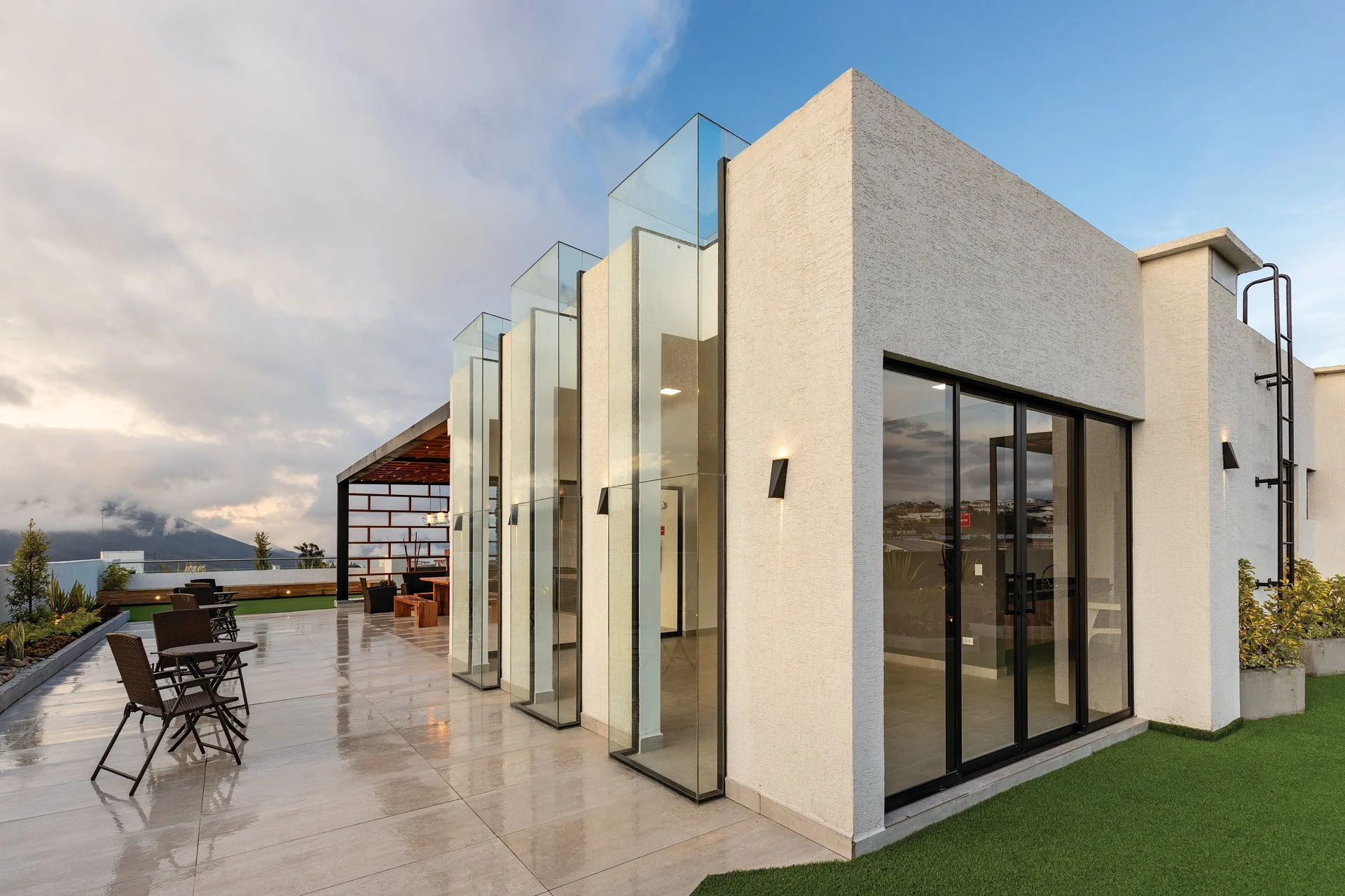 Terraza en un edificio moderno con sillas y plantas, vista al cielo con nubes y montañas al fondo.