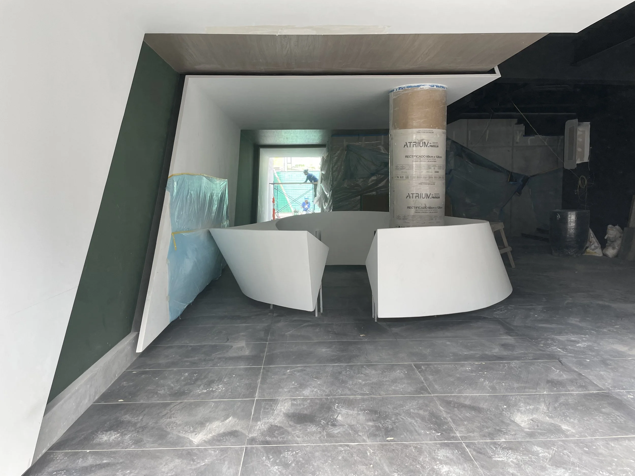 Interior en construcción con paredes en yeso, piso de cerámica gris y una estructura blanca curva en el centro. Se observa una columna envuelta en plástico y un área con materiales de construcción y protección.