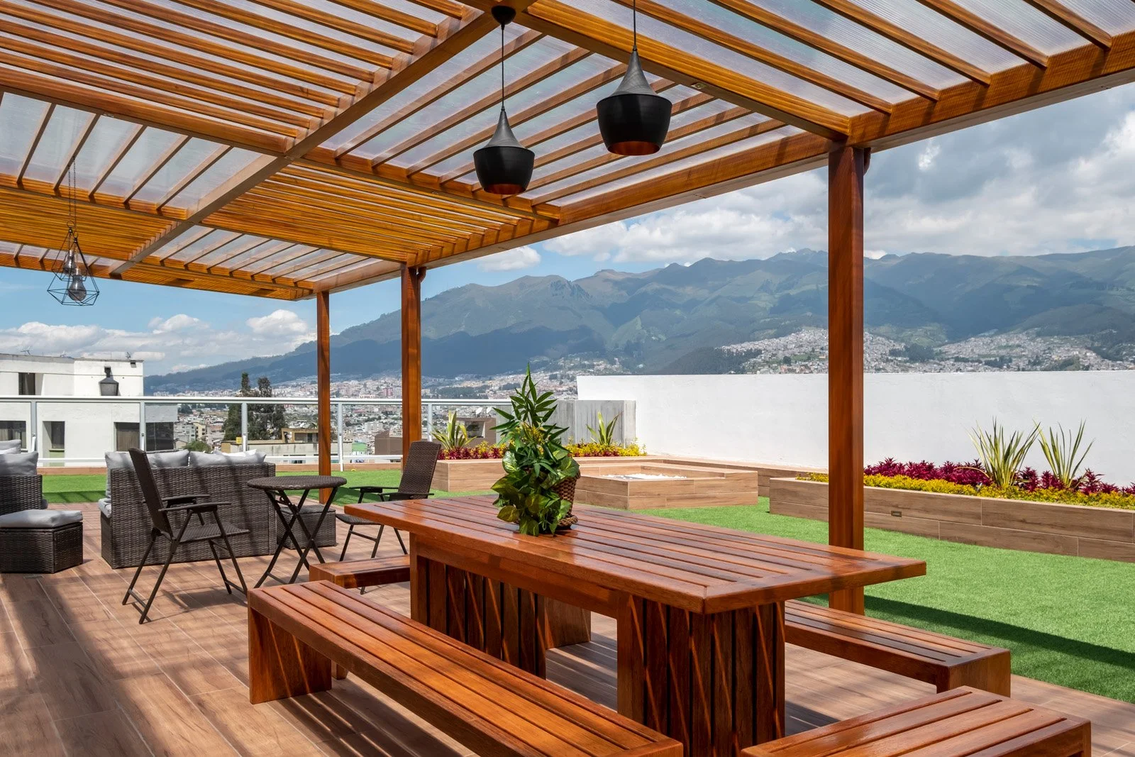 Terraza moderna con pérgola de madera, área de descanso con sillas y mesa, plantas decorativas y vista a las montañas y ciudad en el fondo.