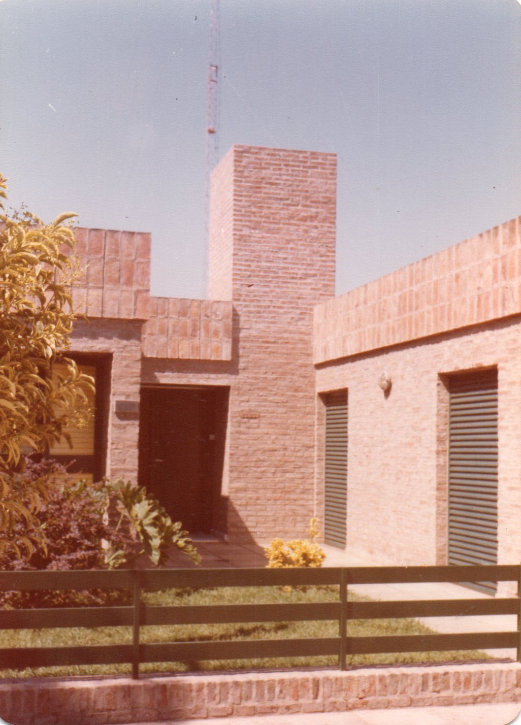 Edificio de ladrillos con ventanas con persianas grises, cerca de jardines y arbustos, con una cerca de madera en primer plano y un poste de radio o televisión en el fondo.