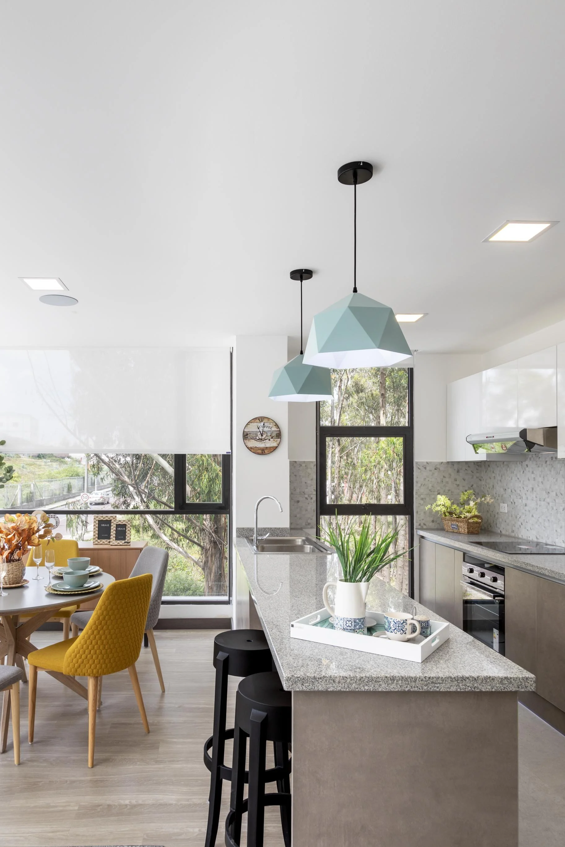 Cocina moderna con isla de granito, mesas y sillas decoradas, lámparas colgantes y ventanas con vista a árboles