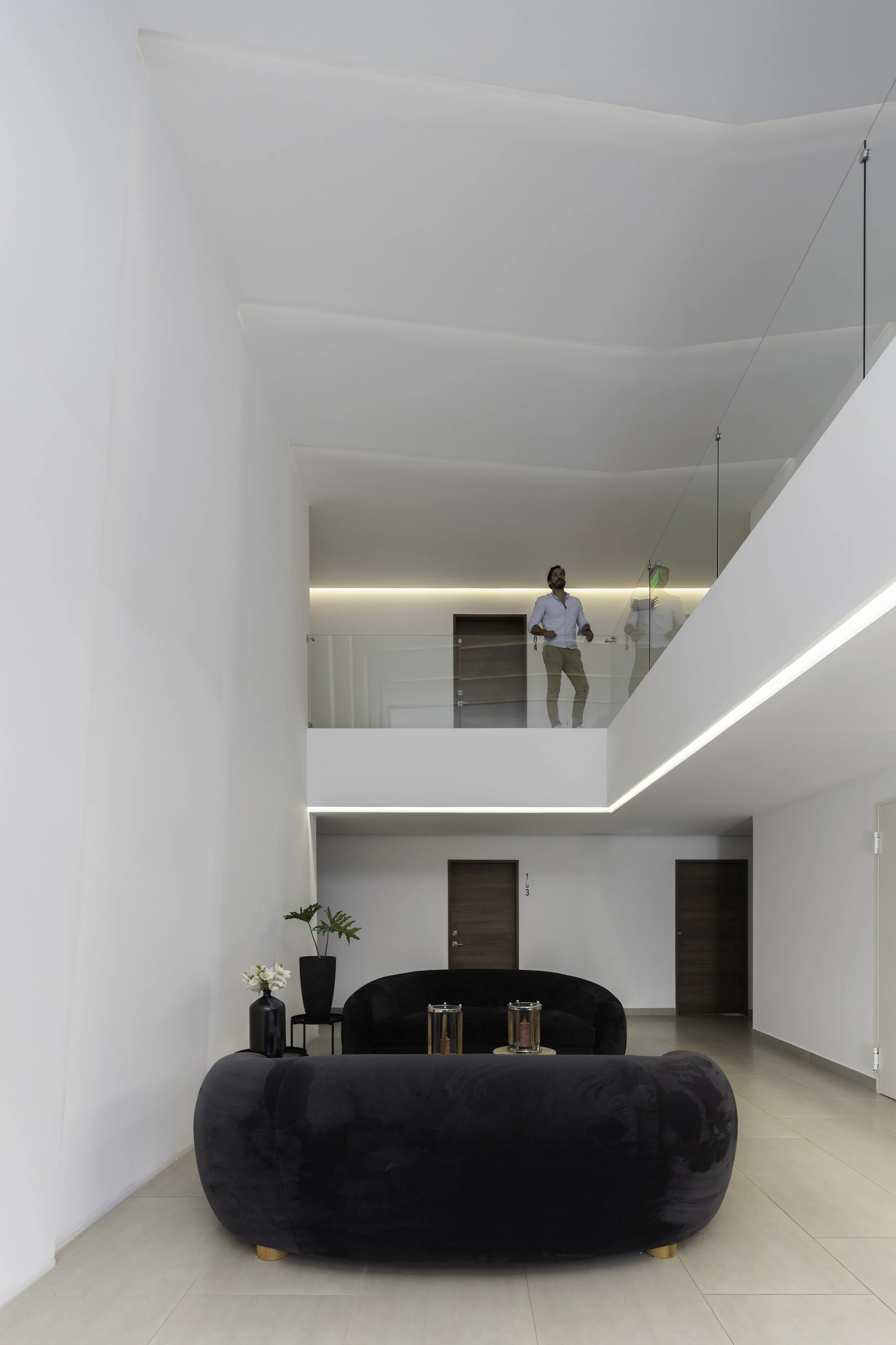 Vista de un interior moderno y minimalista de un edificio con paredes blancas, dos sillones negros y plantas decorativas. En el segundo piso, un hombre con camisa azul claro y pantalones beige observa desde una baranda de vidrio.