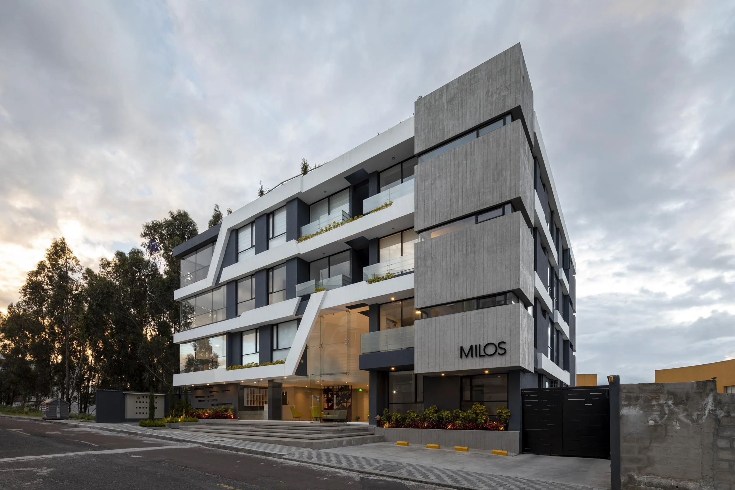 Edificio moderno de varios pisos con ventanas grandes y balcones, cuyo cartel dice 'Milos', en un fondo de cielo nublado y árboles a su lado.