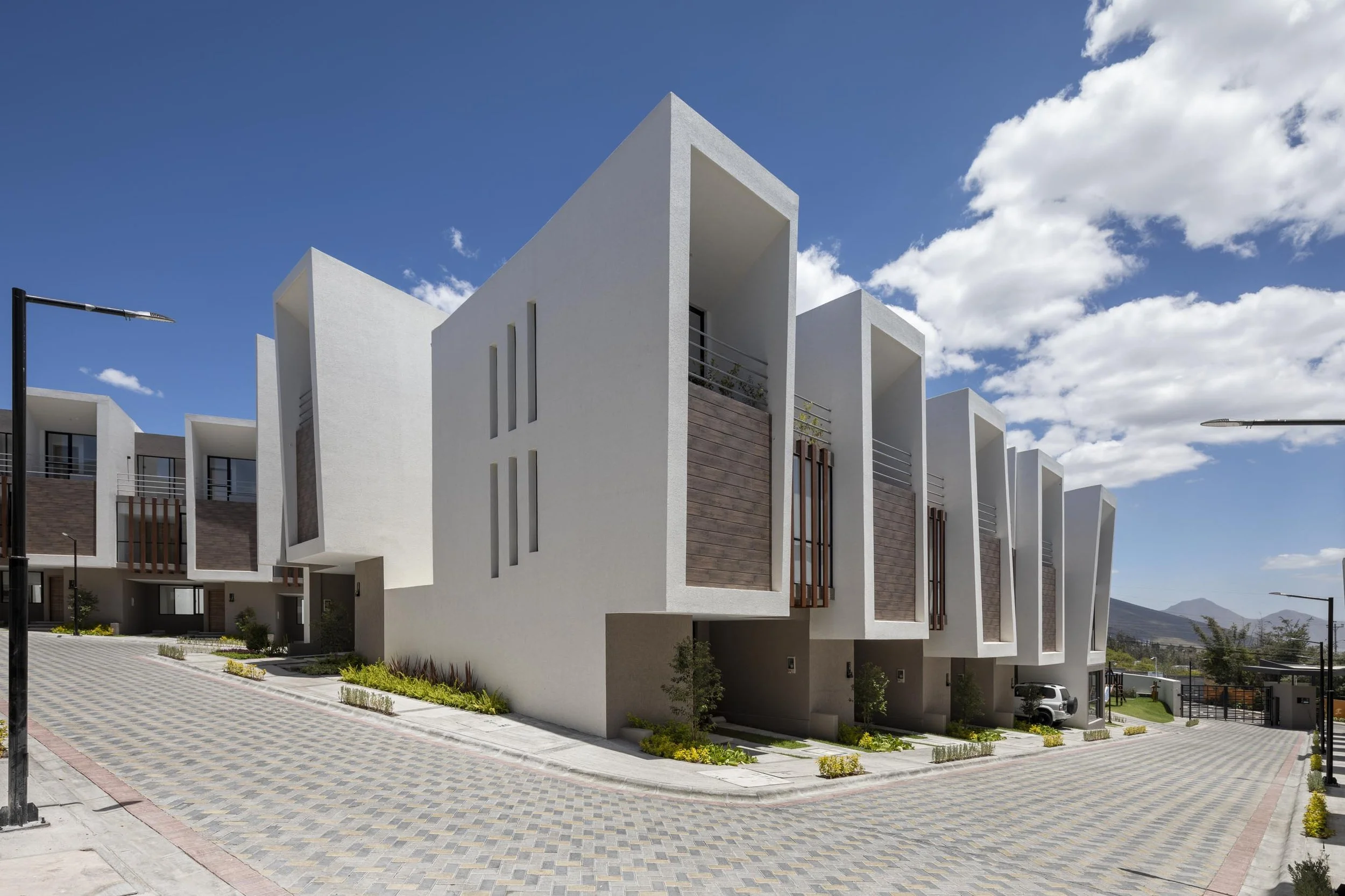Conjunto de casas modernas blancas con ventanas estrechas, en un vecindario con calles pavimentadas, árboles pequeños y un cielo azul con nubes.