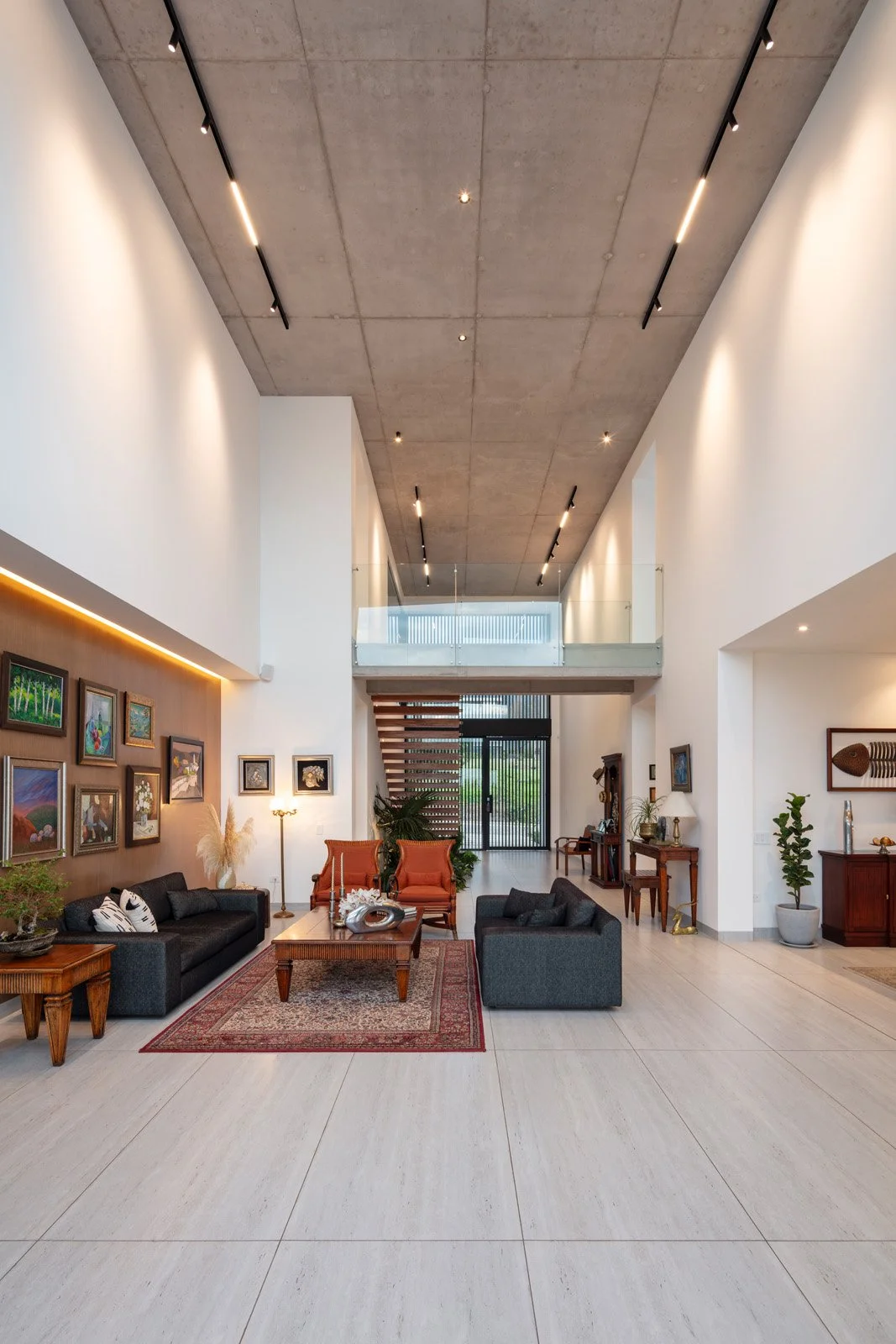 Sala de estar moderna con techo alto de concreto, muebles de estilo clásico con sofás y sillas, paredes decoradas con cuadros, y entrada con escaleras de madera a un segundo nivel.