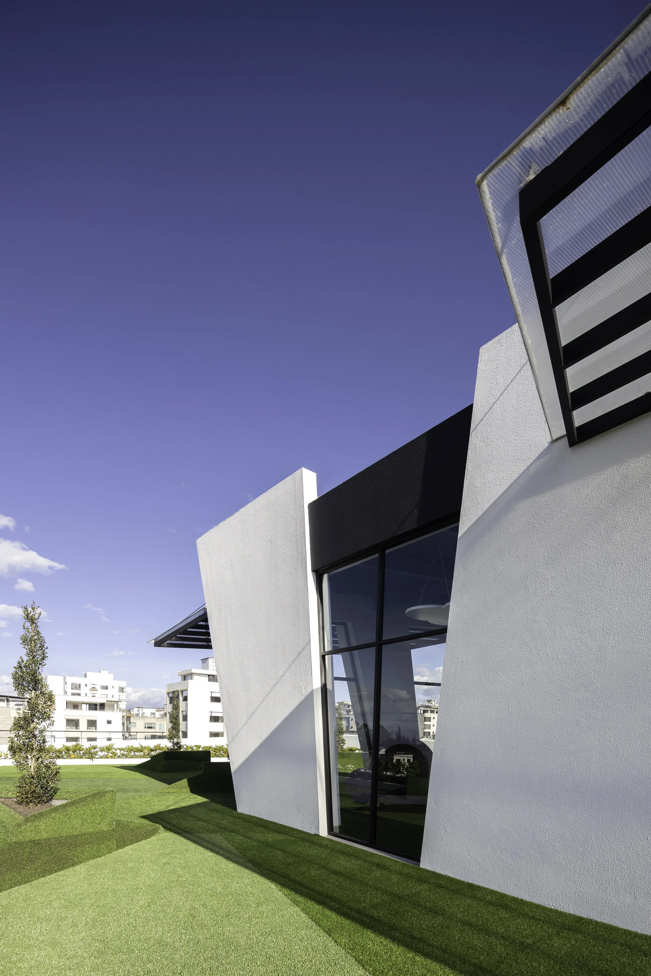 Vista de un edificio moderno con paredes blancas y ventanas de cristal, rodeado de césped verde y un cielo azul despejado.