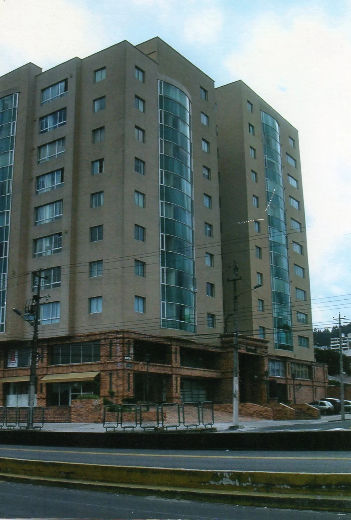 Edificio de varios pisos con fachada de ladrillo y ventanas de vidrio, situado junto a una calle con barreras y postes de luz.