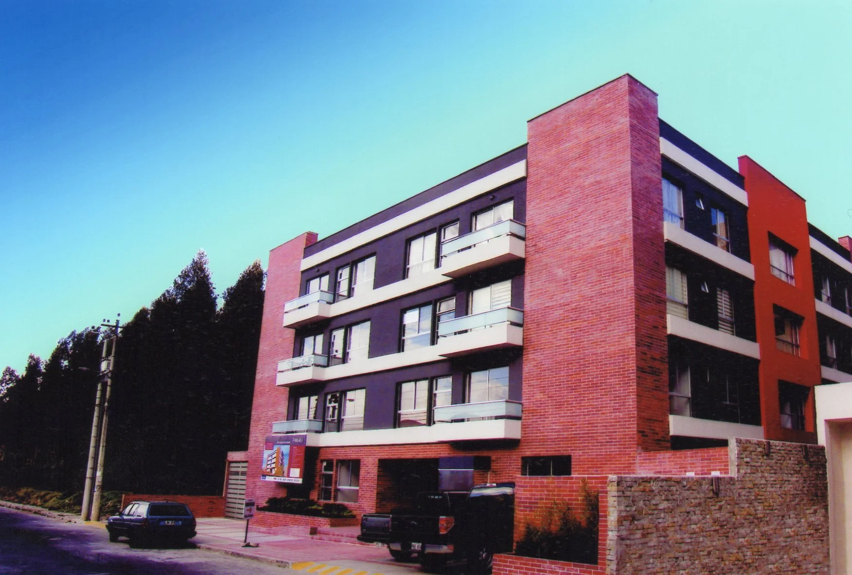 Edificio moderno de varios pisos con fachadas de ladrillos rojos y paredes negras, balcones blancos y ventanas grandes, frente a una calle con automóviles y señalización de tránsito.