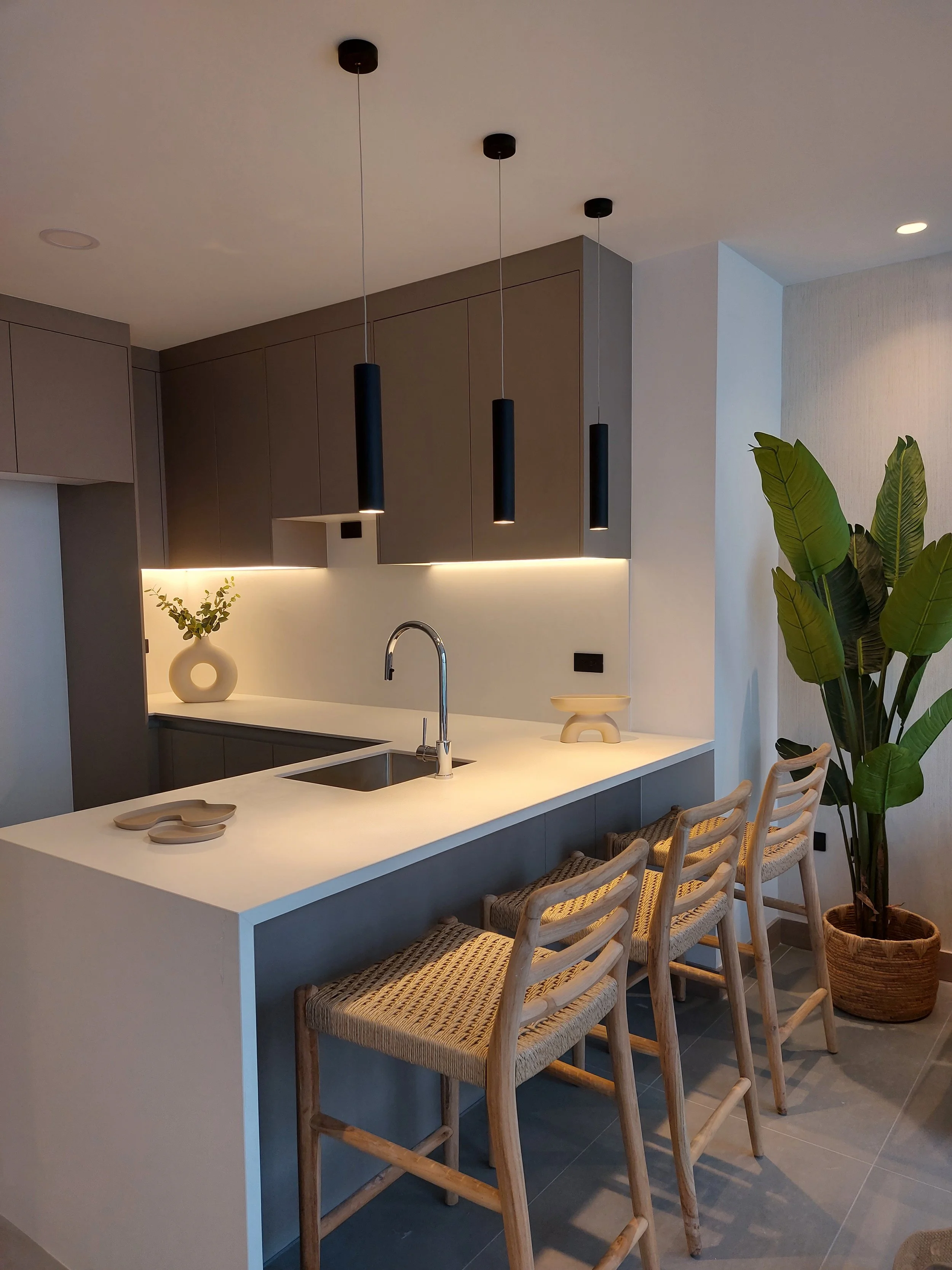 Cocina moderna con una isla, tres sillas de madera, gabinetes grises, tres lámparas colgantes negras, y plantas decorativas.