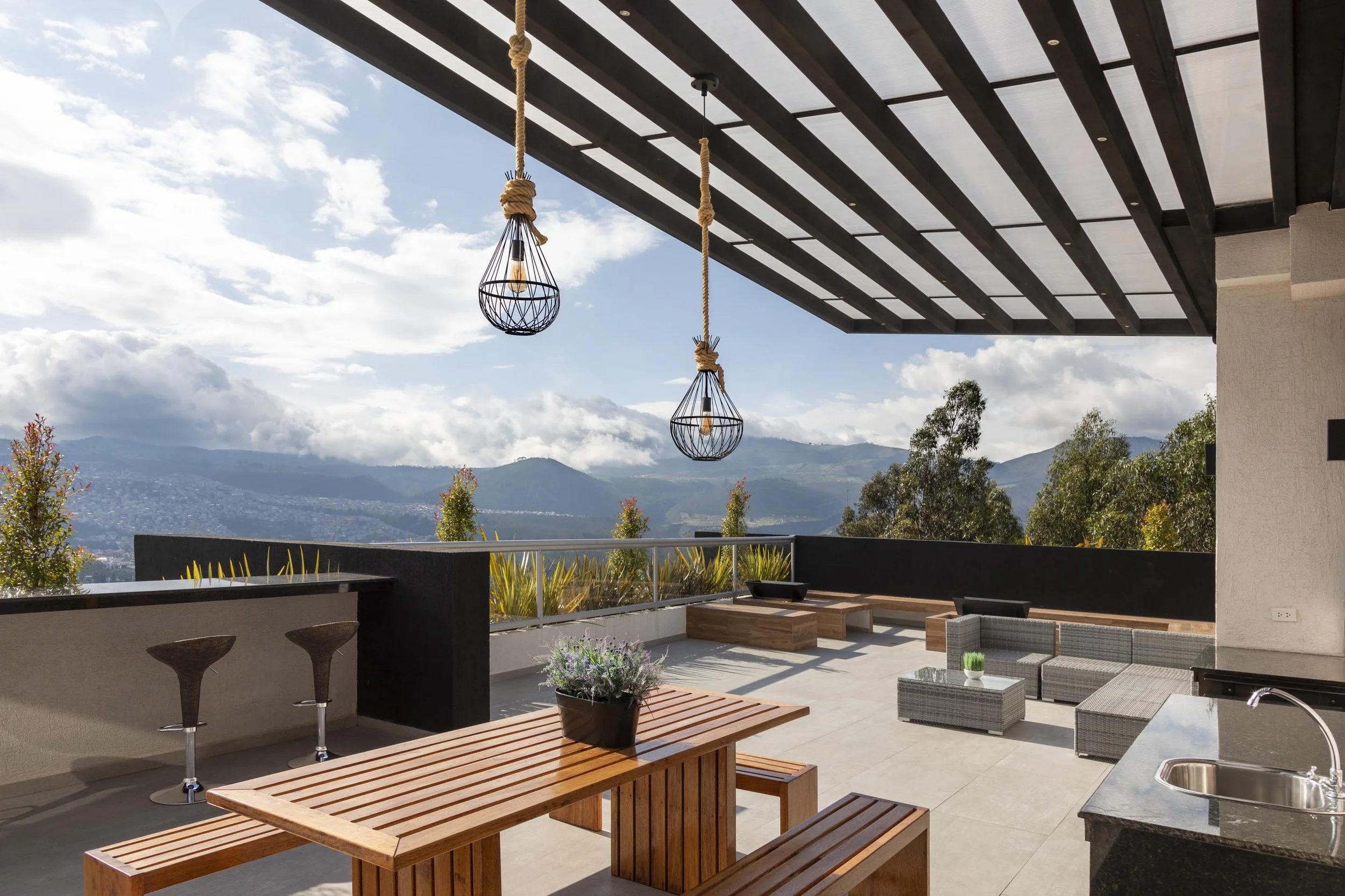 Terraza con mesas de madera, sillones de ratán, iluminación de lámparas colgantes y vista de montañas y árboles al fondo.