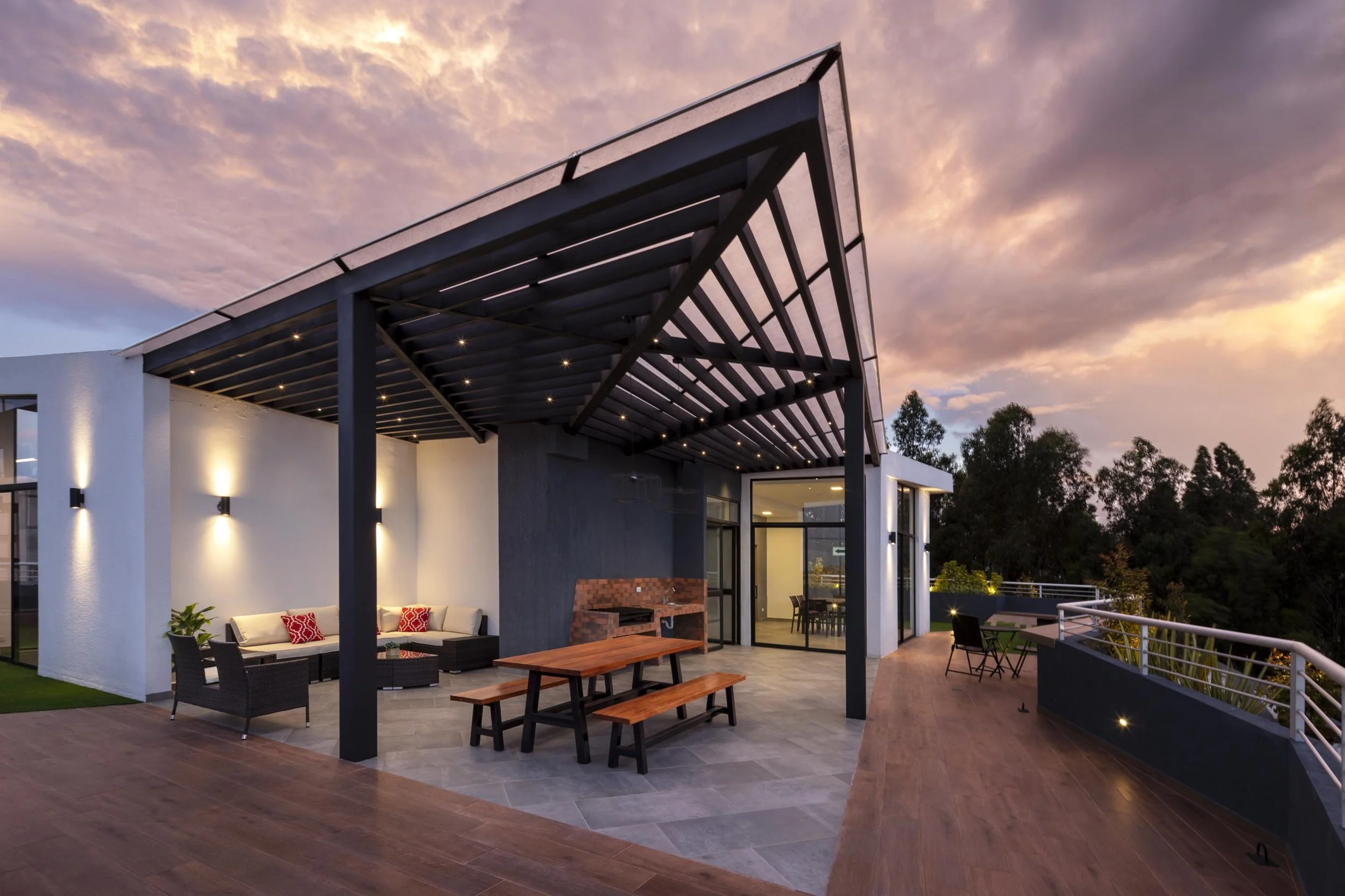 Terraza moderna con sillones, mesa de picnic de madera, barbacoa de ladrillo y vista al atardecer con árboles al fondo.