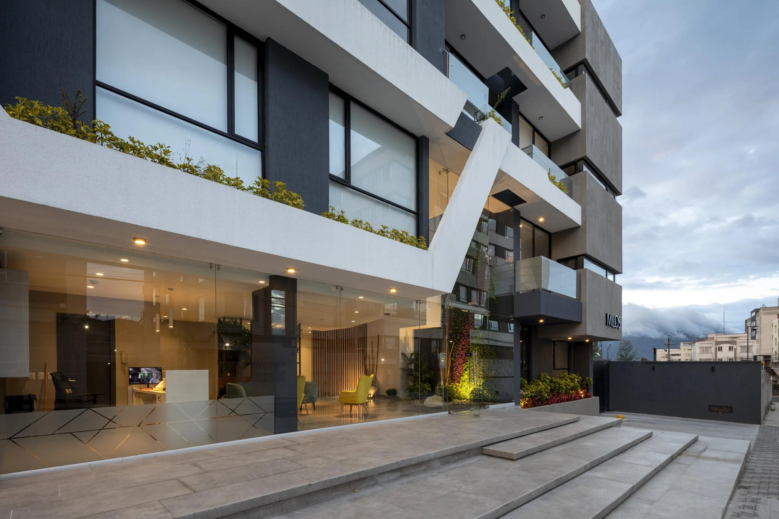 Edificio moderno de varios pisos con fachada de cristal y balcones con jardineras, entrada principal con escaleras y áreas verdes iluminadas.