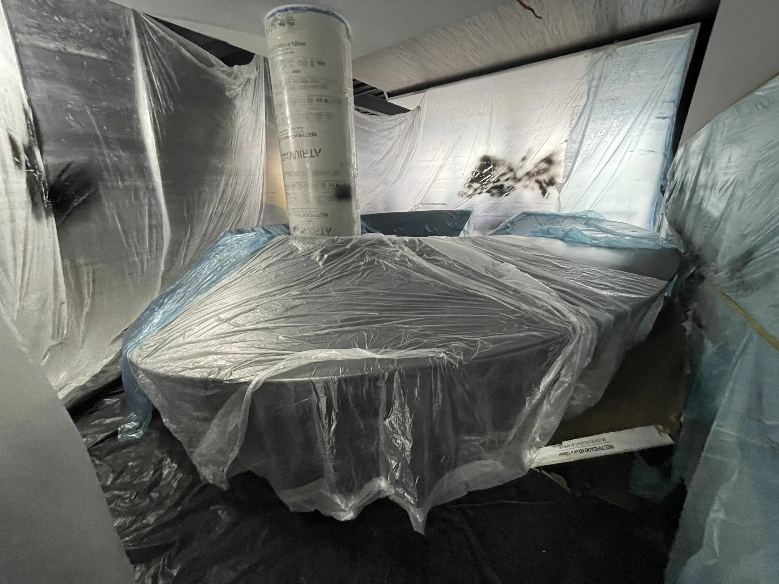 Una habitación en construcción o renovación, con muebles cubiertos con plástico transparente y una tubería frente a la cama, paredes parcialmente cubiertas con protector de plástico para evitar polvo y escombros.