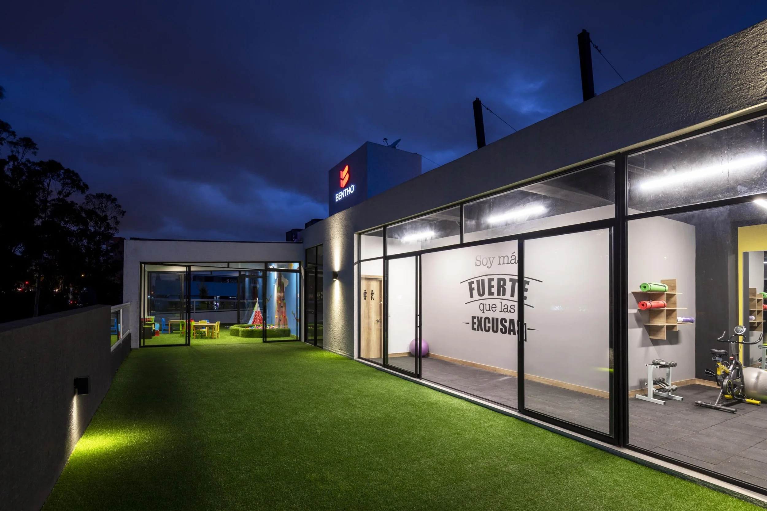 Vista exterior de un gimnasio con ventanas de cristal, área de juegos para niños, y espacio de ejercicio con pelotas y bicicletas, en la noche con cielo oscuro.
