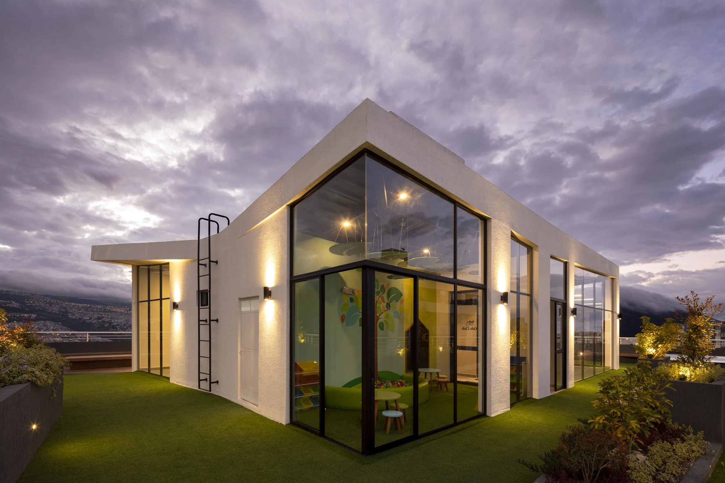 Edificio moderno de dos pisos con paredes blancas y grandes ventanas de vidrio, iluminado con luces exteriores, en un entorno urbano con colinas y nublado en el cielo.