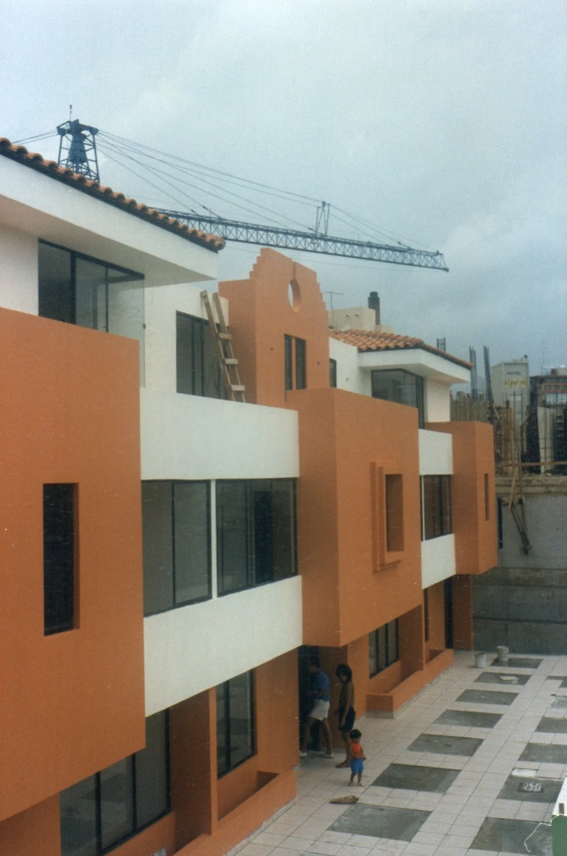 Edificio en construcción con detalles en color naranja y blanco, mostrador de azulejos en el suelo, personas y una grúa de construcción en el fondo.