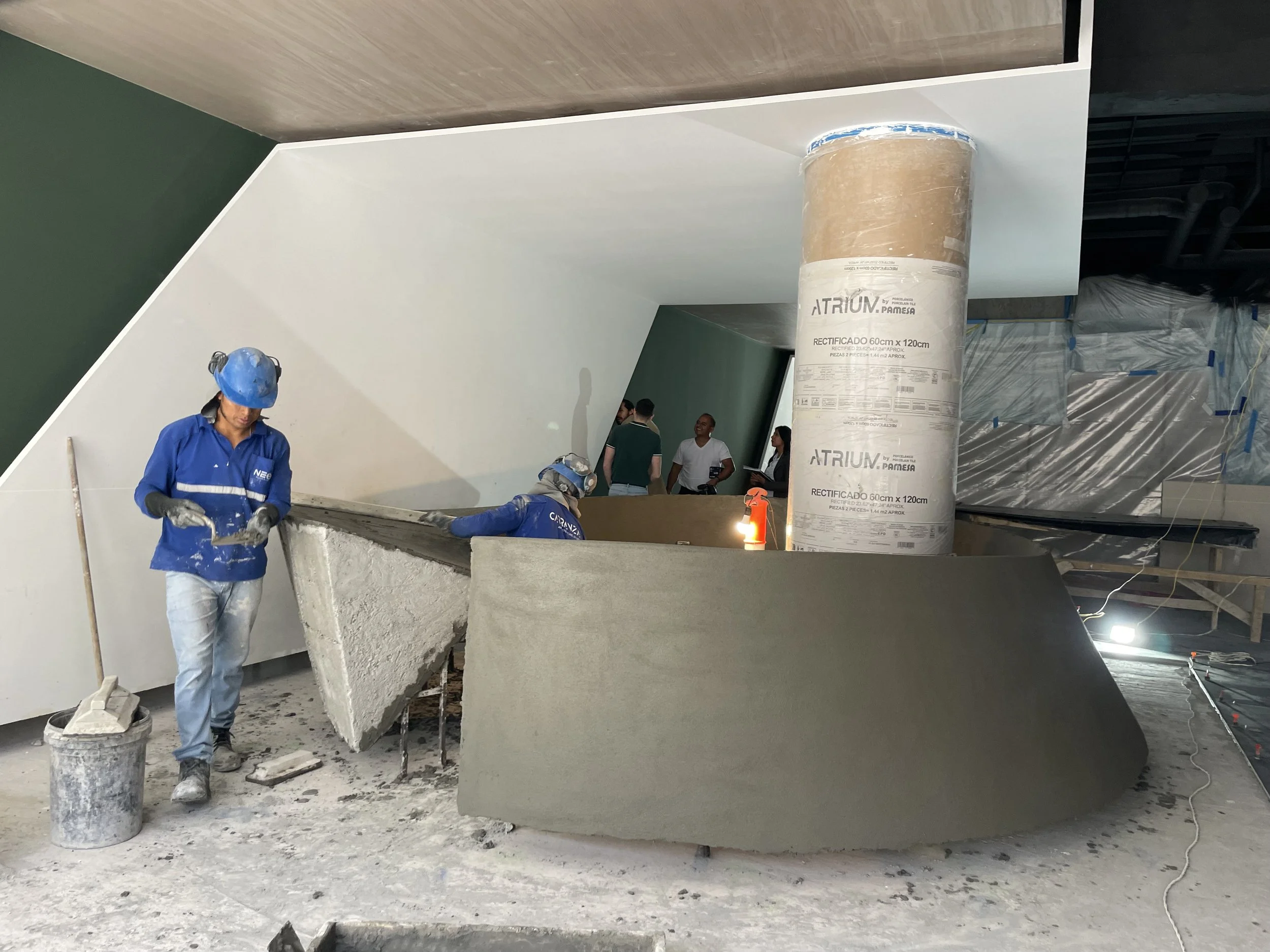 Trabajadores en construcción, uno con casco azul y guantes, trabajando en una estructura de concreto y paredes en proceso de acabado.
