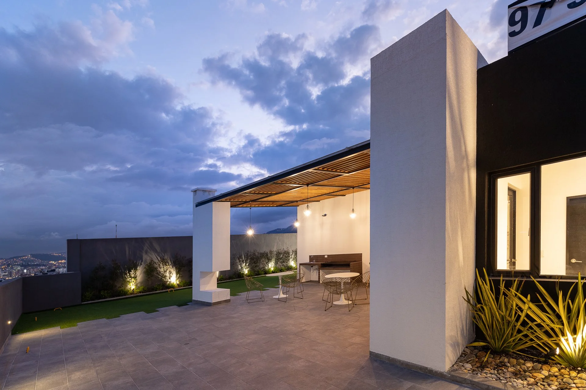 Terraza moderna con mesa y sillas, iluminación exterior y vista urbana al atardecer.