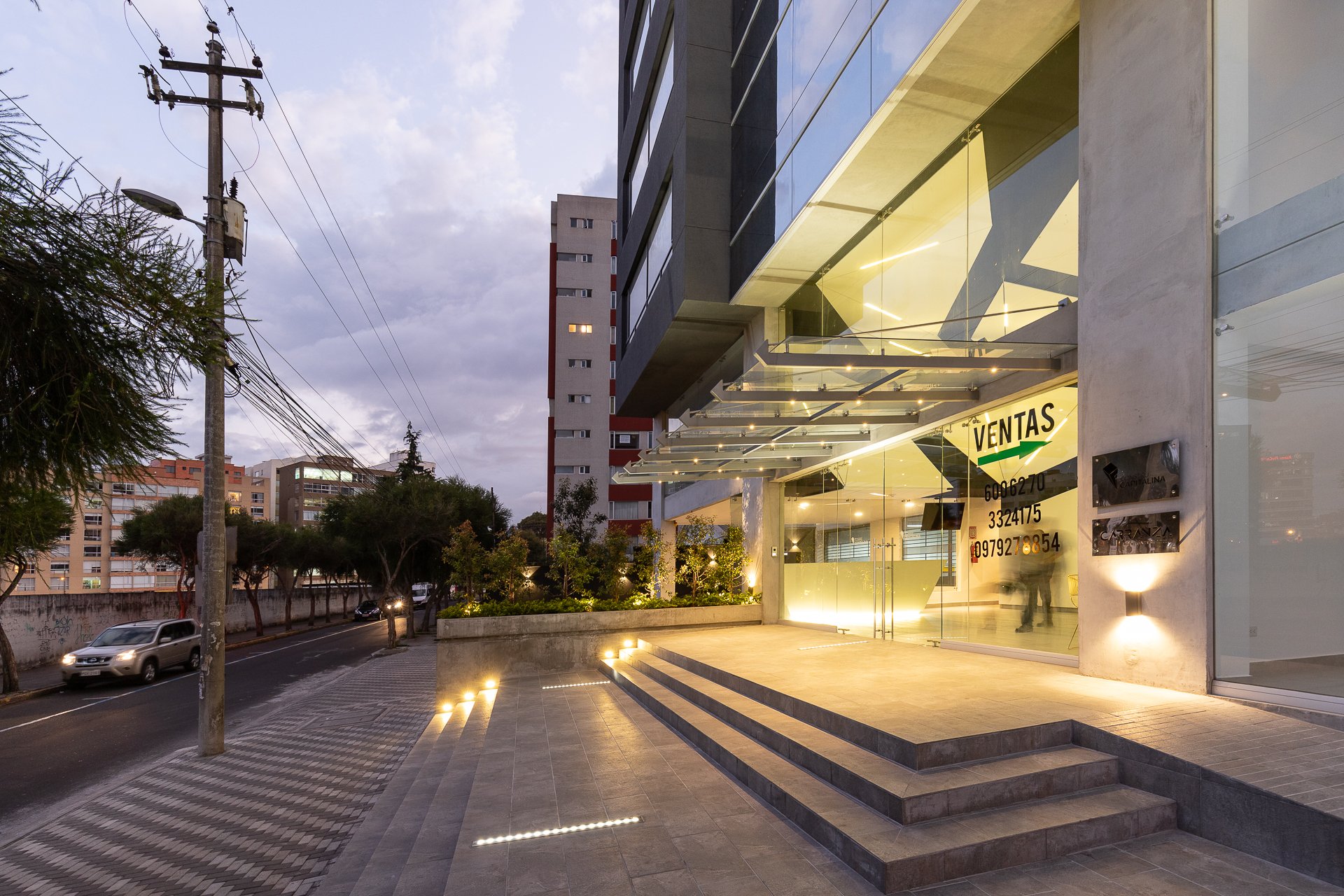 Fachada de un edificio con área de ventas, con escalones y paredes de cristal, iluminada durante el atardecer en una ciudad.
