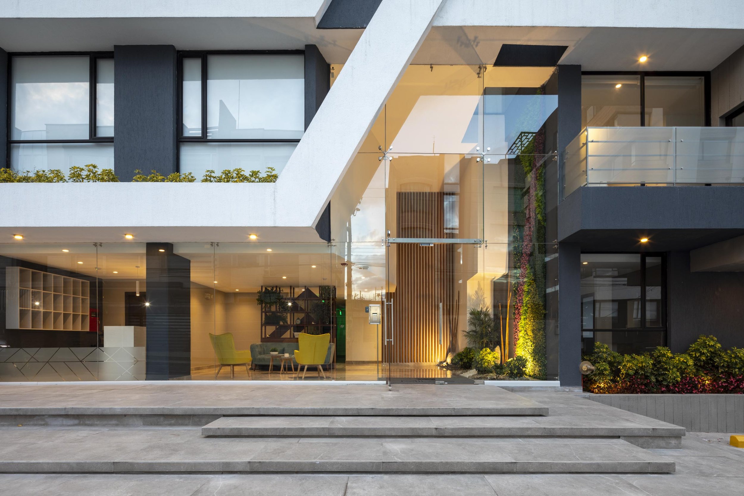 Fachada moderna de edificio con entrada de cristal, espacio en interior con sillones verdes y plantas decorativas, y escalones en primer plano.