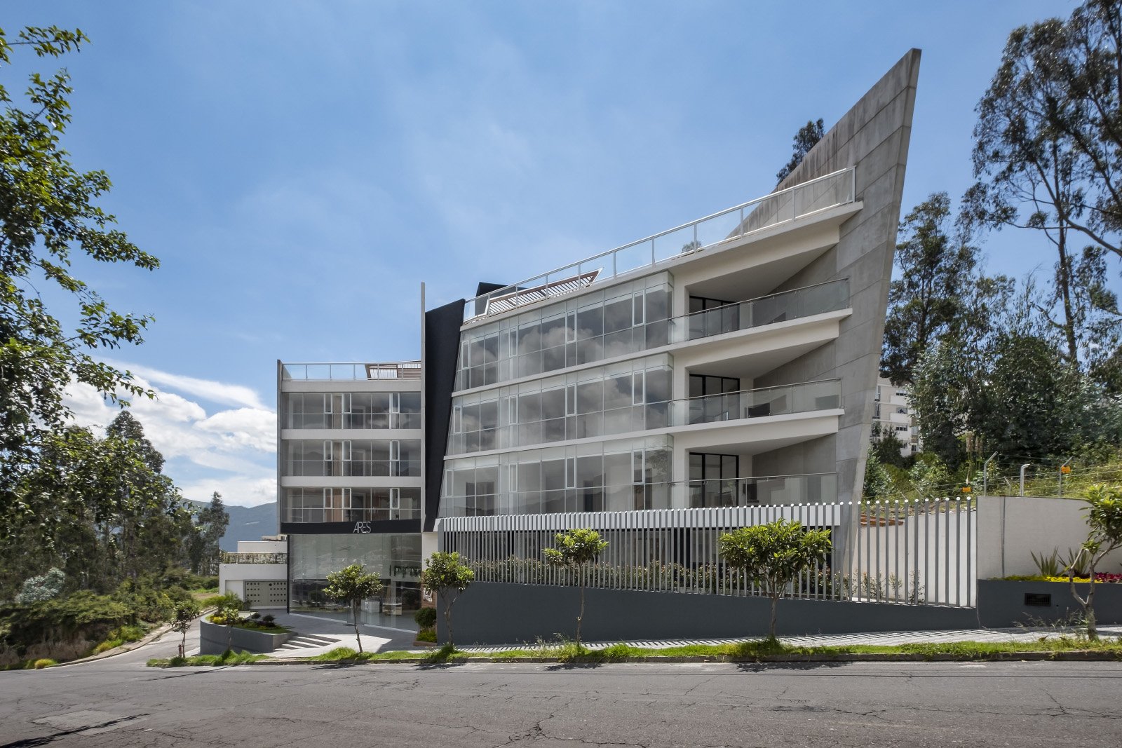 Edificio moderno de varios pisos con estructuras de concreto y amplios ventanales de vidrio, rodeado de árboles y una calle en primer plano, bajo un cielo parcialmente nublado.