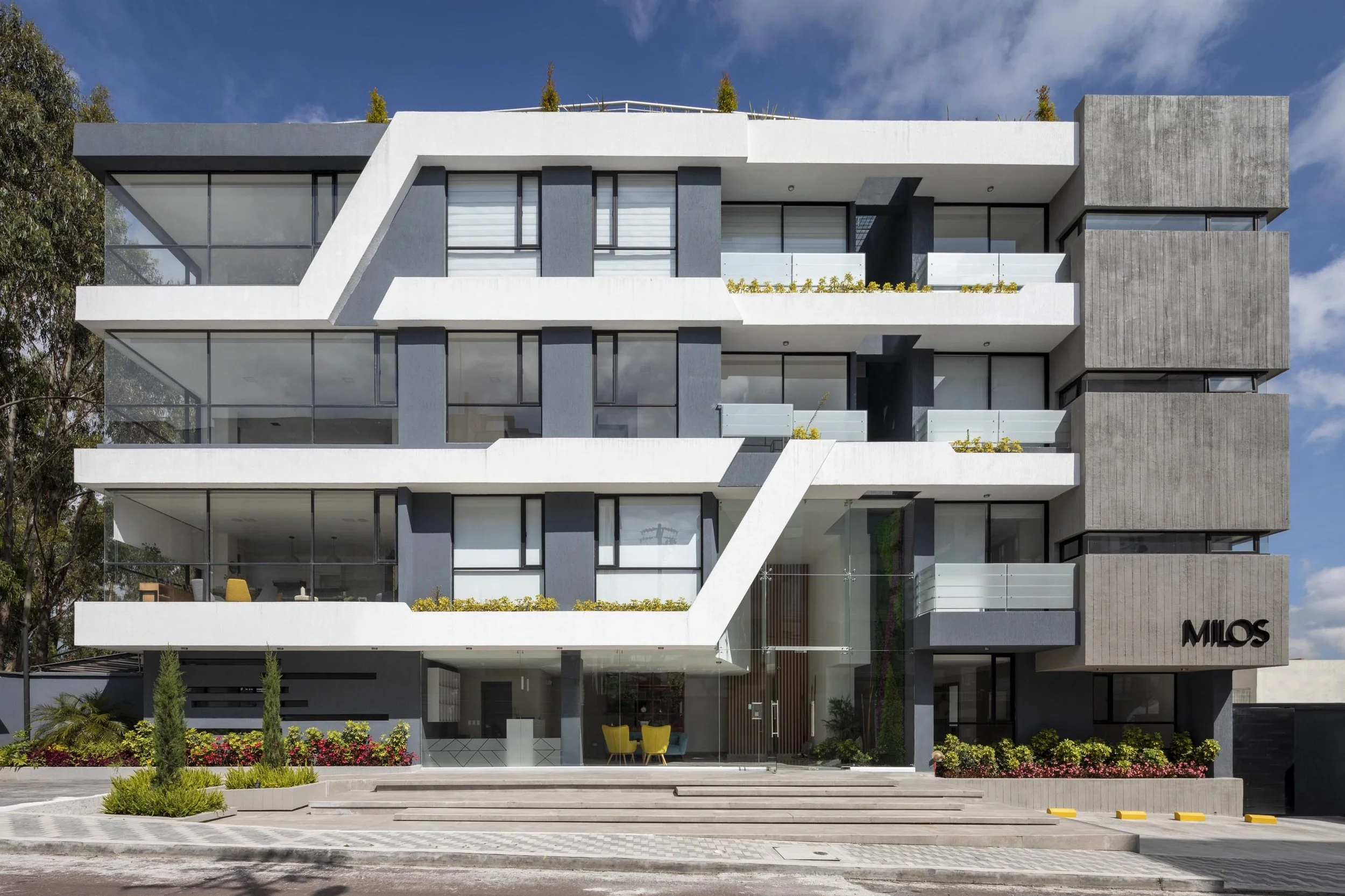 Edificio de apartamentos moderno con fachada de concreto gris, balcones con barandales de vidrio y plantas en las jardineras, entrada con escalones y señalización que dice 'Milos', árboles y cielo con nubes al fondo.