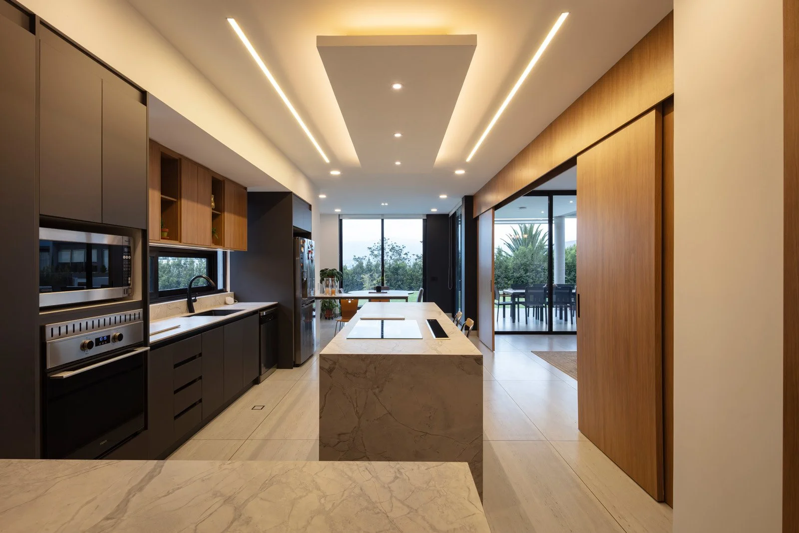 Cocina moderna con encimera de mármol, gabinetes oscuros y ventanas grandes que muestran un jardín exterior