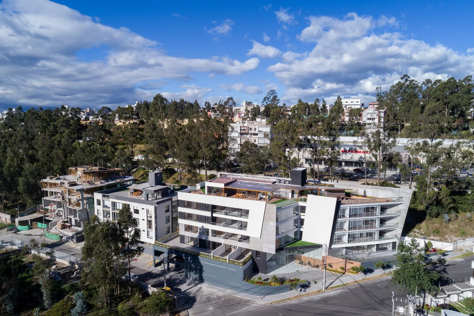 Complejo de edificios modernos en una zona urbana rodeada de árboles y áreas en construcción, bajo un cielo parcialmente nublado.