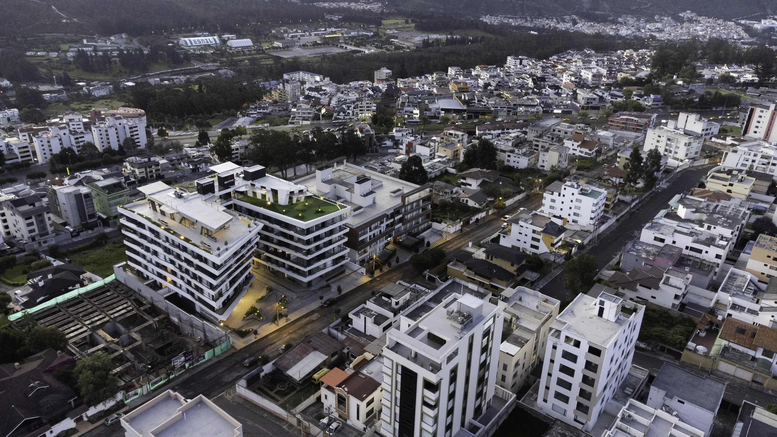 Vista aérea de una ciudad moderna con edificios de diferentes alturas, incluyendo edificios residenciales y comerciales, durante el atardecer.